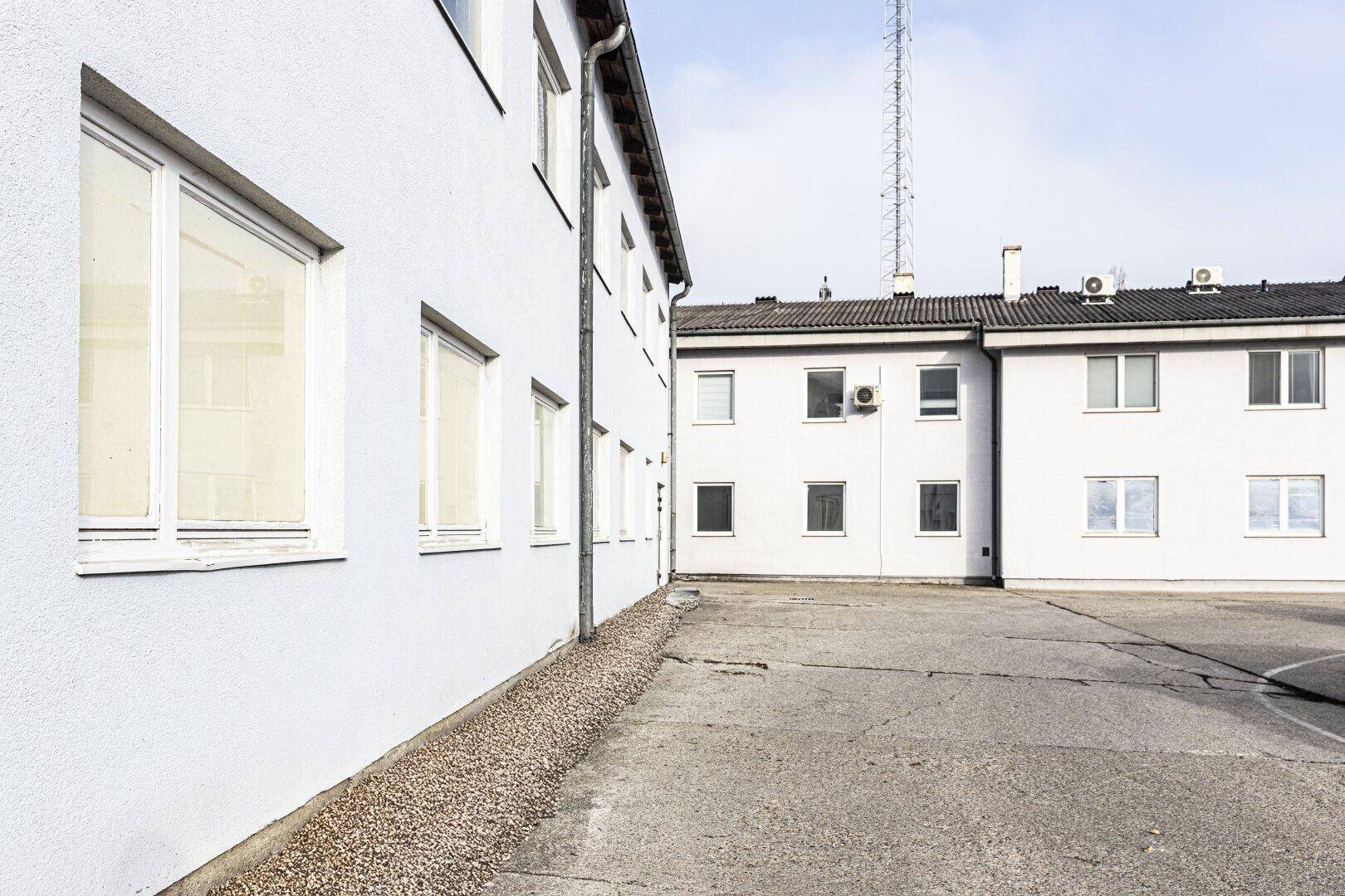 Weiße Gebäudefassade mit mehreren Fenstern und einem Kiesstreifen am Sockel, daneben ein gepflasterter Hofbereich mit weiteren Gebäuden im Hintergrund.