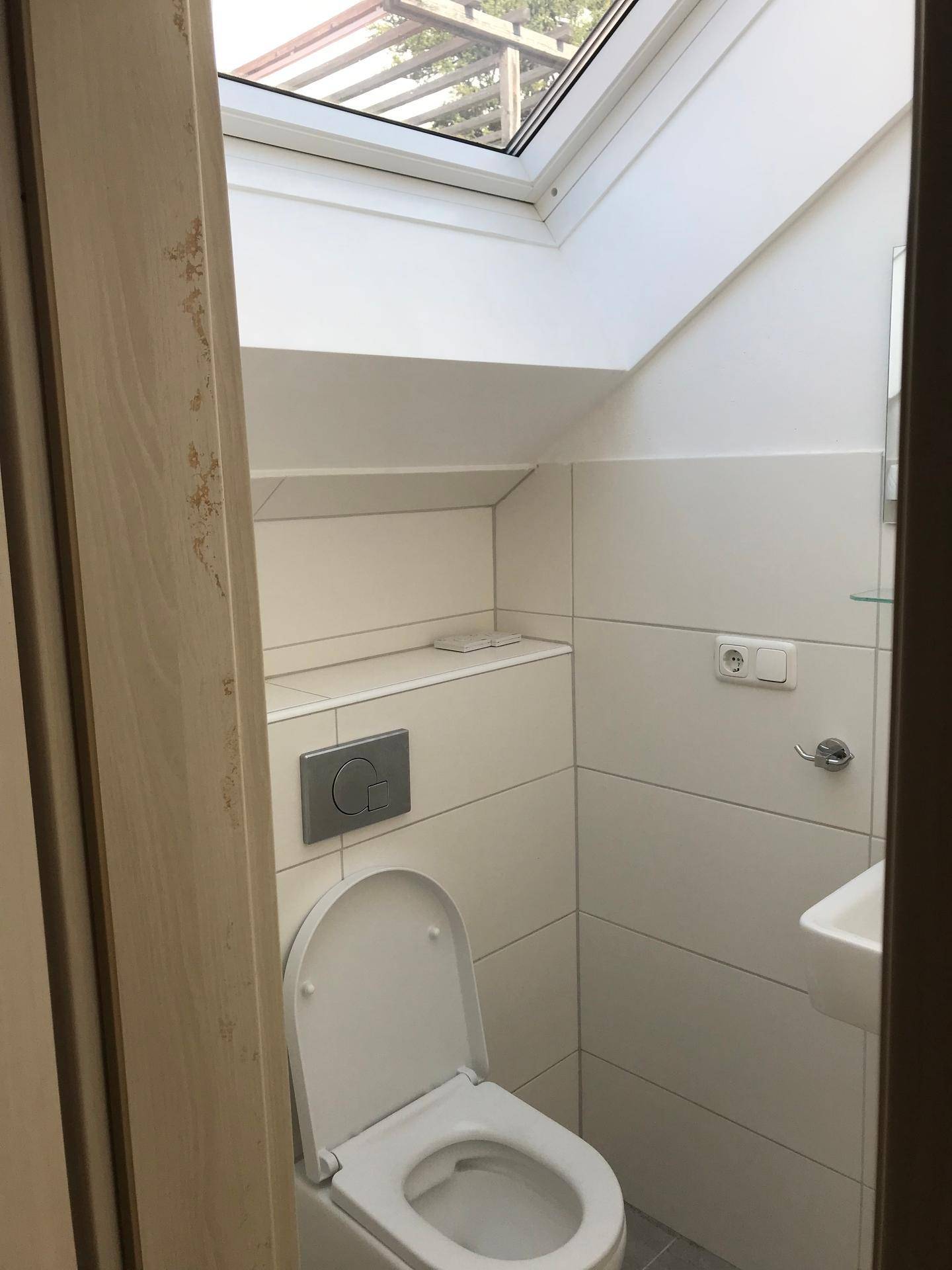 Helles WC mit wandhängender Toilette, kleinem Waschbecken und einem Dachfenster für Tageslicht.