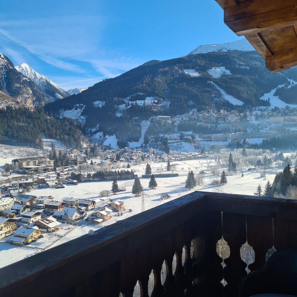 Blick vom Holzbalkon auf eine verschneite Alpenlandschaft mit Dorf und Bergen.