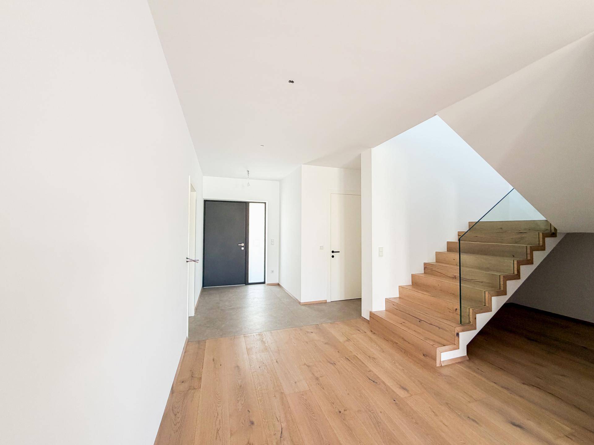 Moderner Eingangsbereich mit Holzboden, offener Treppe und dunkler Eingangstür.