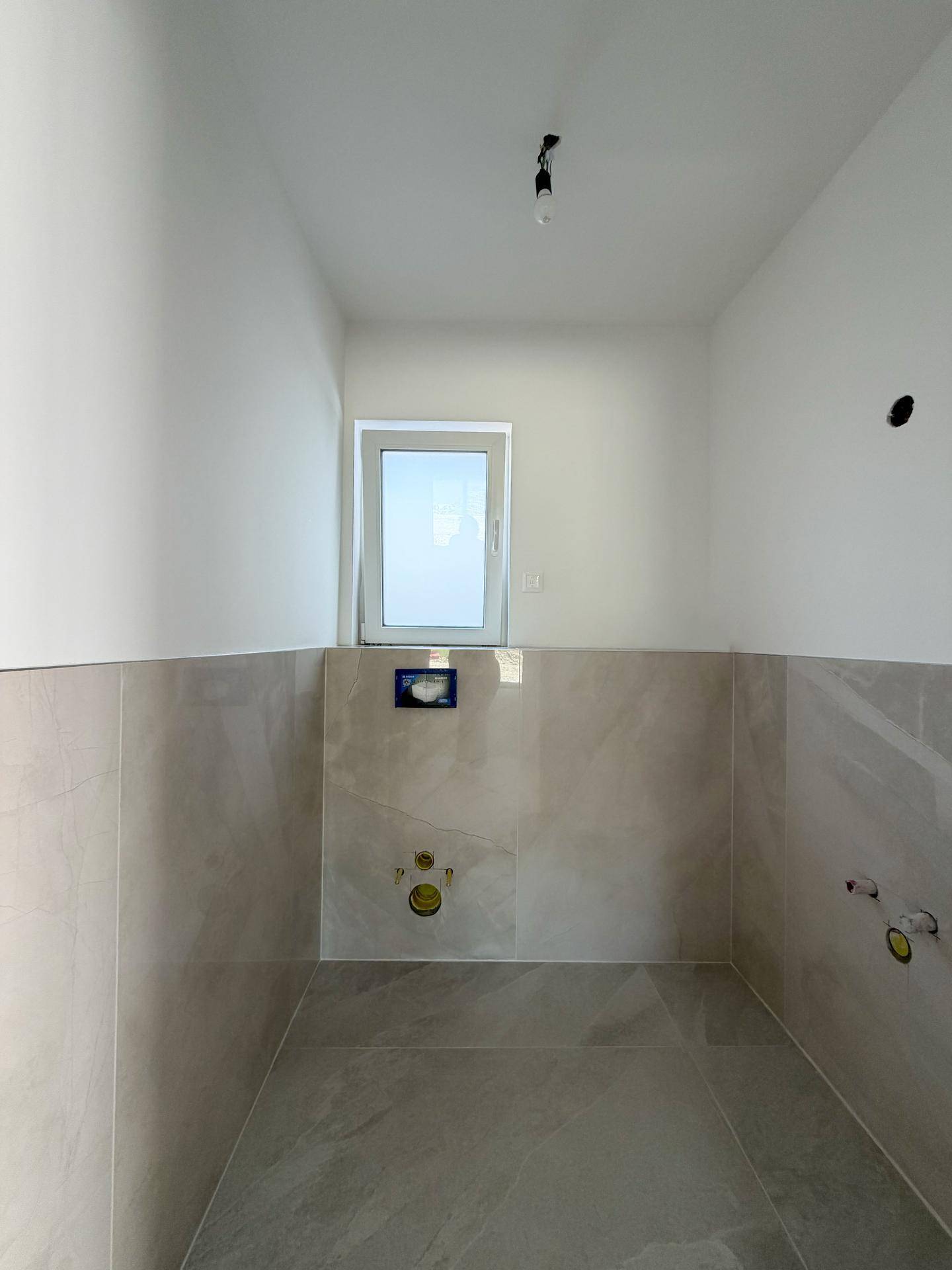 Kleiner Raum im Rohbau mit Fliesenboden, Fenster und Vorbereitung für ein Wand-WC.