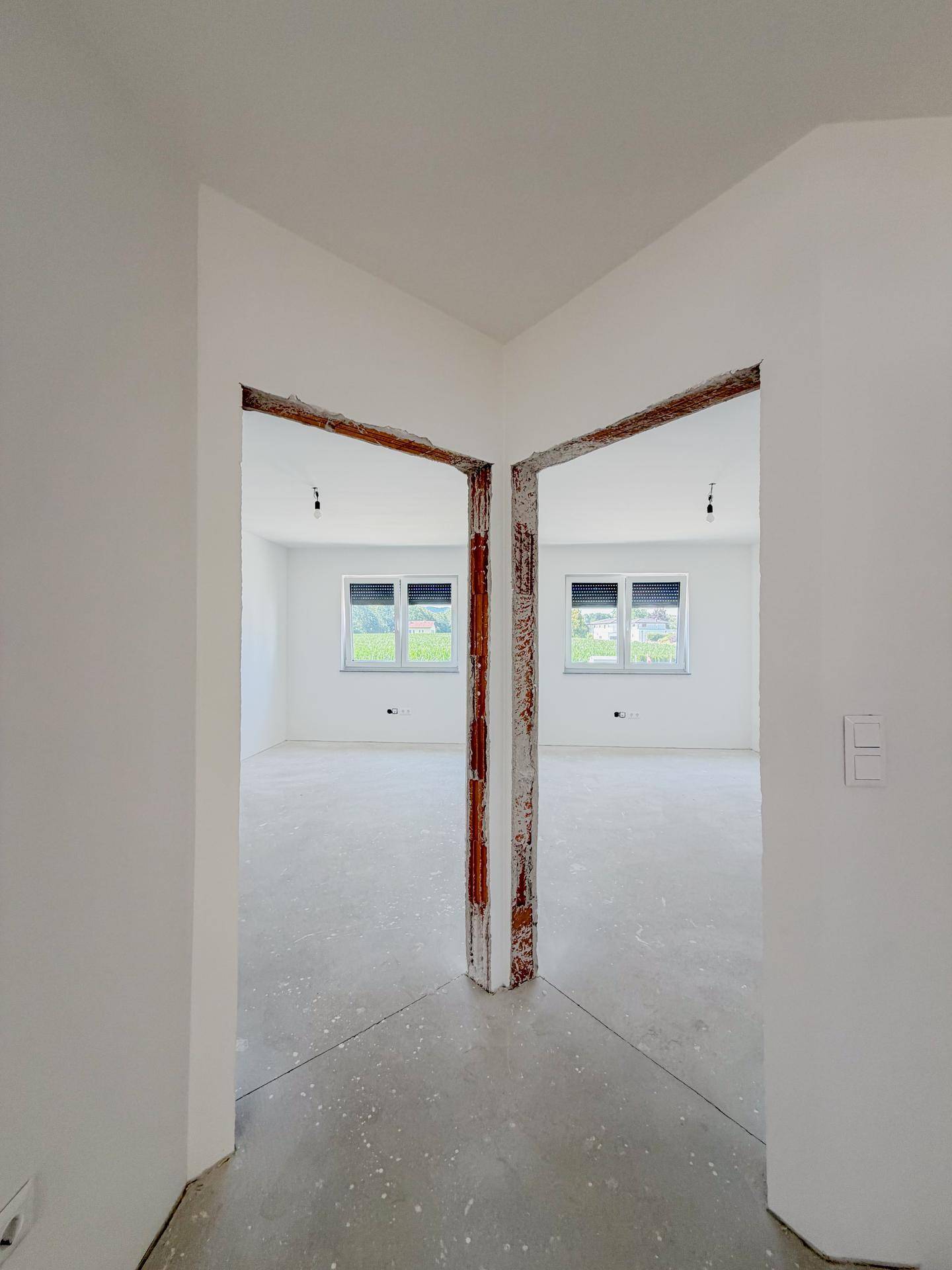 Raum im Rohbau mit Betonboden, zwei Fenstern und Blick ins Grüne.