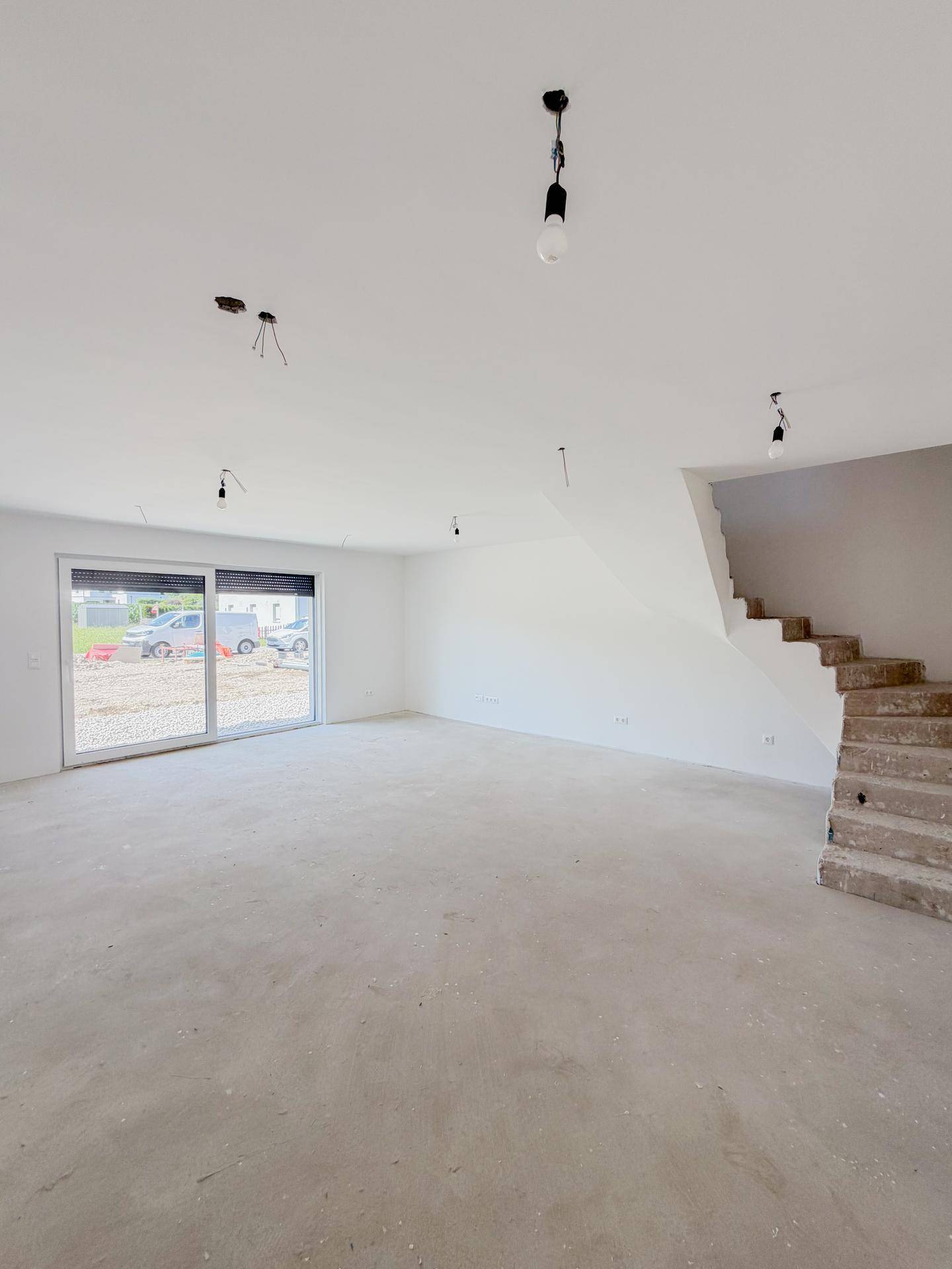 Großes Wohnzimmer im Rohbauzustand mit Betonboden und viel Tageslicht durch die Terrassentür.