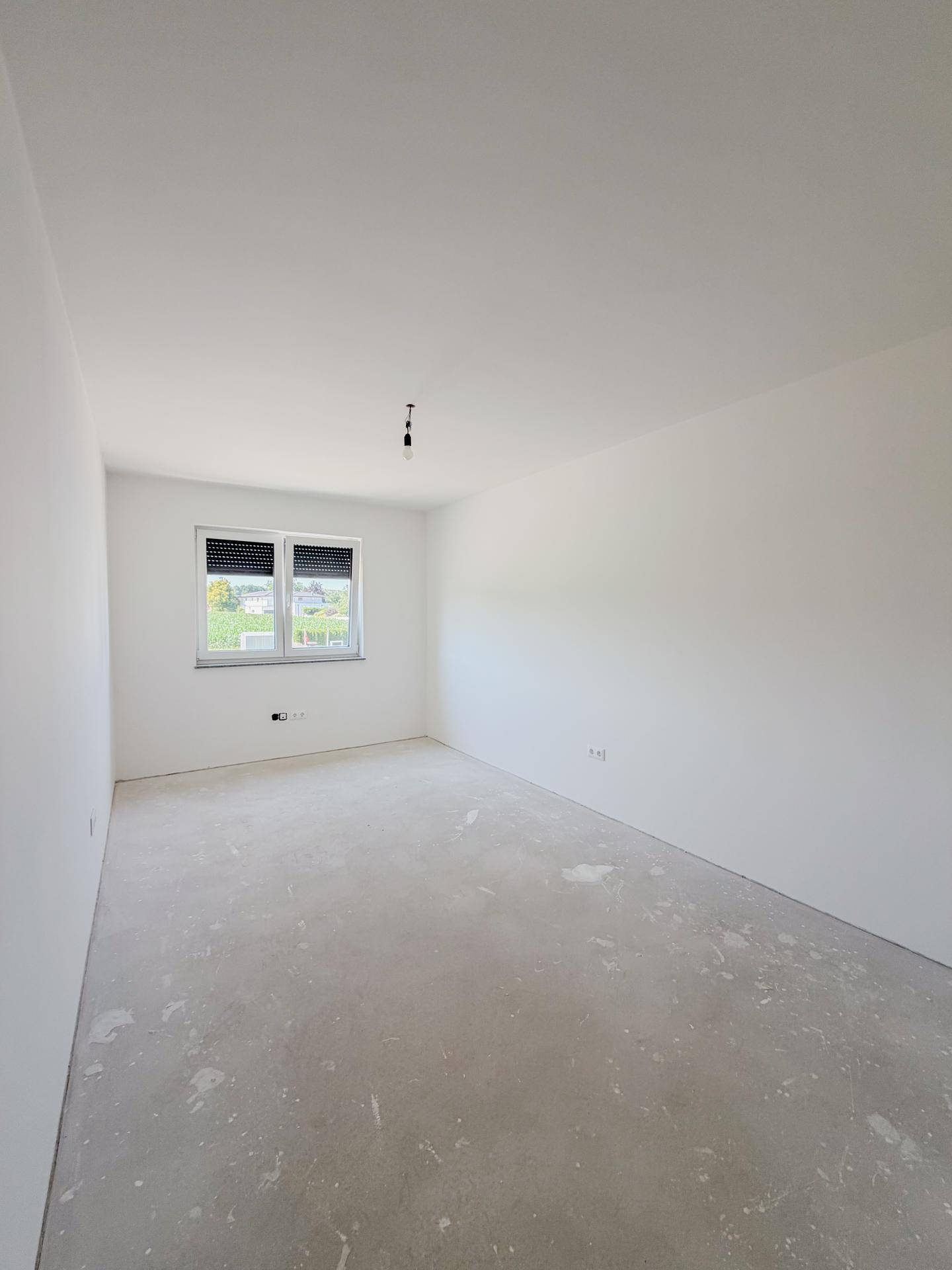 Schlafzimmer im Rohbau mit Betonboden, weiß gestrichenen Wänden und einem Fenster.