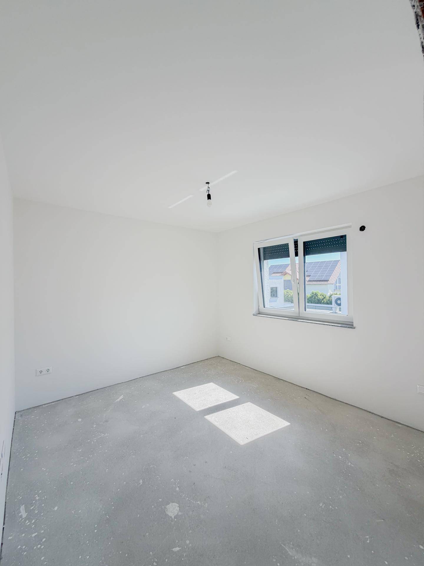 Helles Schlafzimmer im Rohbau mit Betonboden und einem Fenster, das viel Tageslicht hereinlässt.