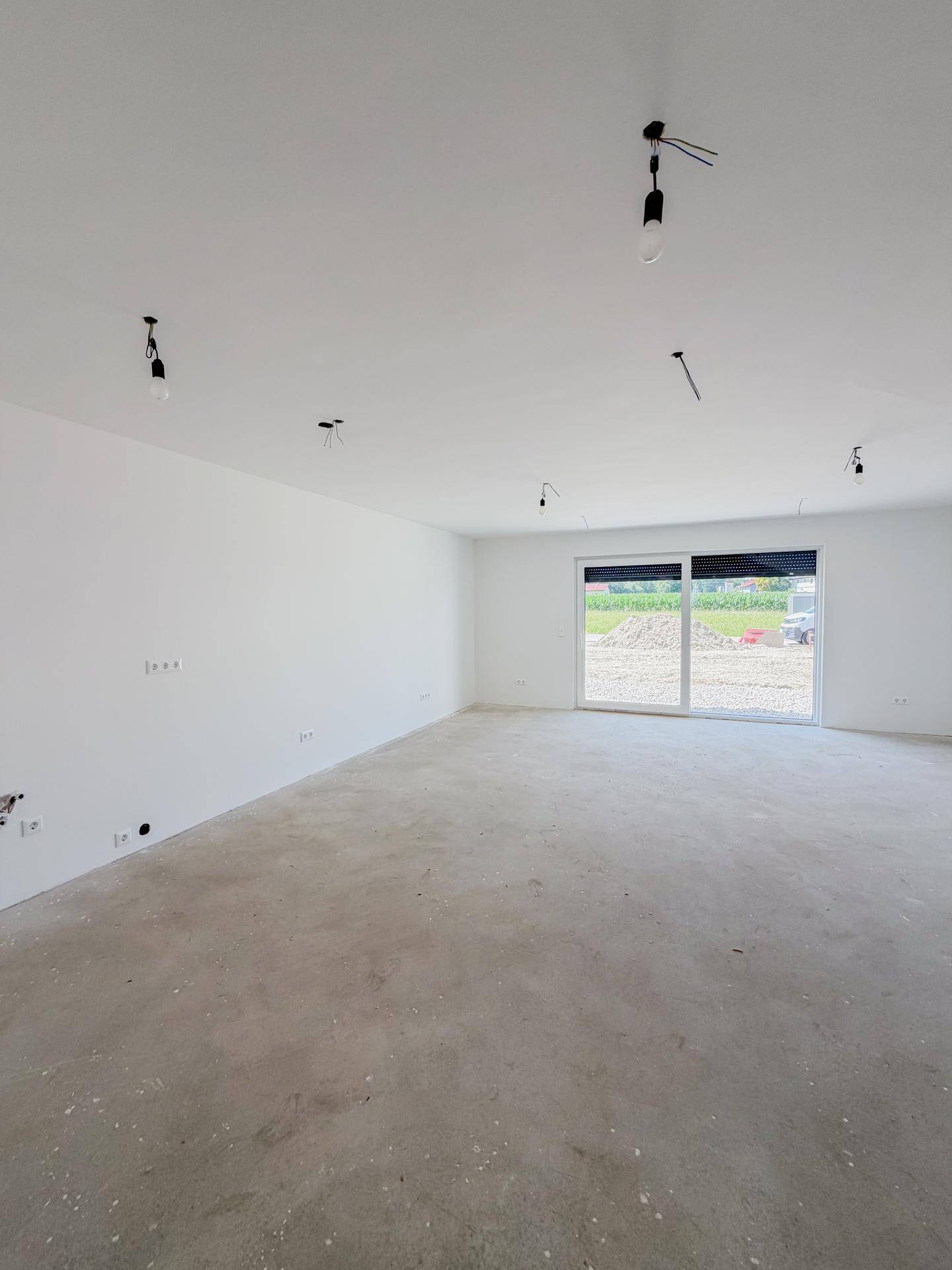Weitläufiges Wohnzimmer im Rohbau mit Betonboden und großer Fensterfront zum Außenbereich.