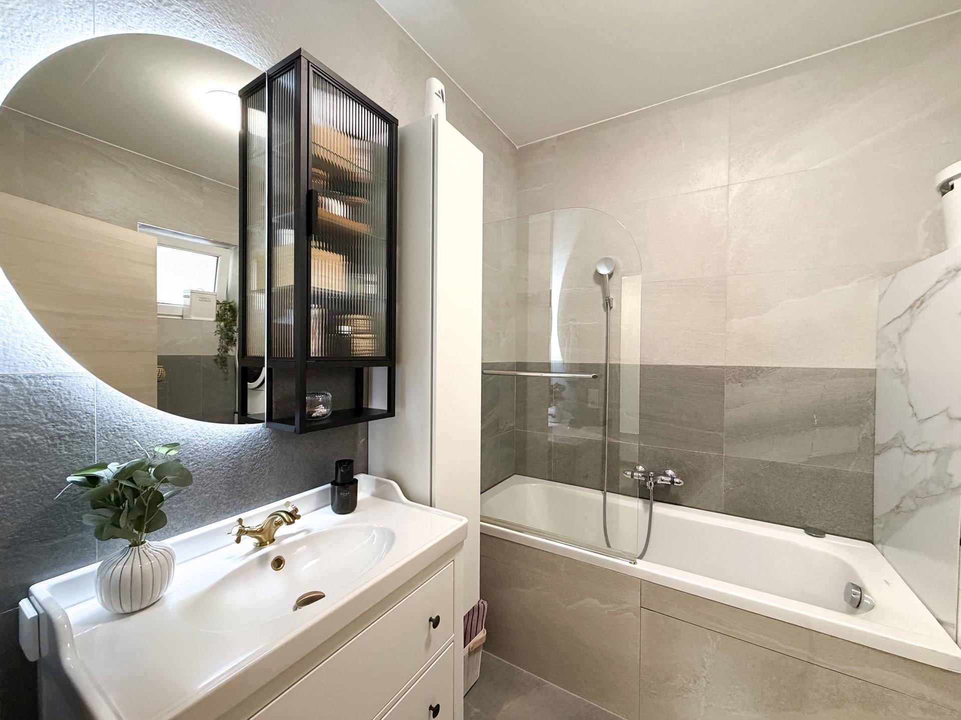 Modernes Badezimmer mit Badewanne, beleuchtetem Spiegel und stilvollen grauen Fliesen.