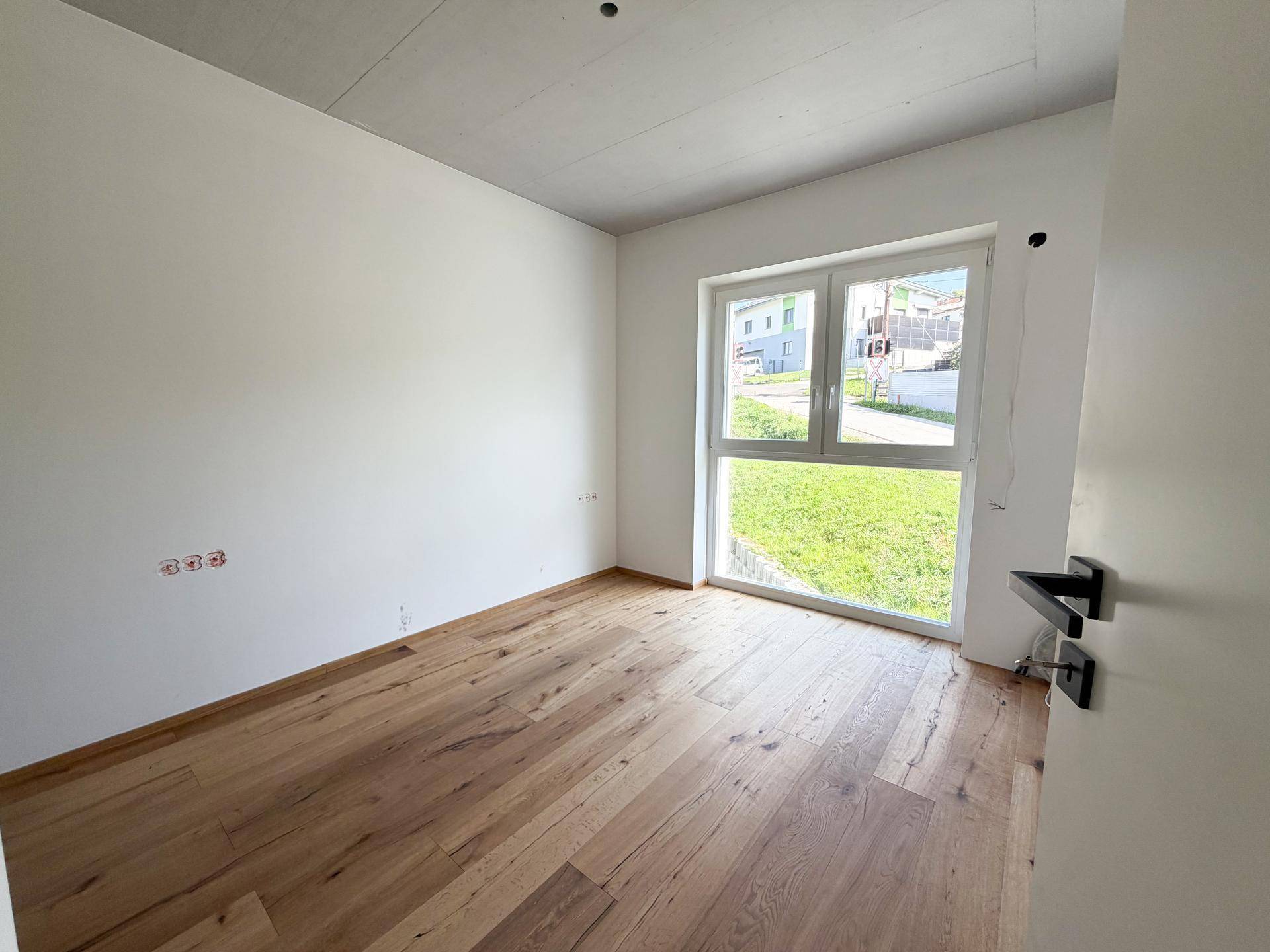 Helles Zimmer mit Holzboden, weiß gestrichenen Wänden und großem Fenster mit Grünblick.