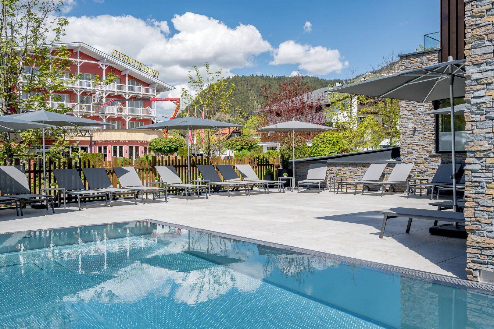 Sonniger Außenbereich mit großem Pool, Liegestühlen und Schirmen, umgeben von grüner Landschaft und Gebäuden.