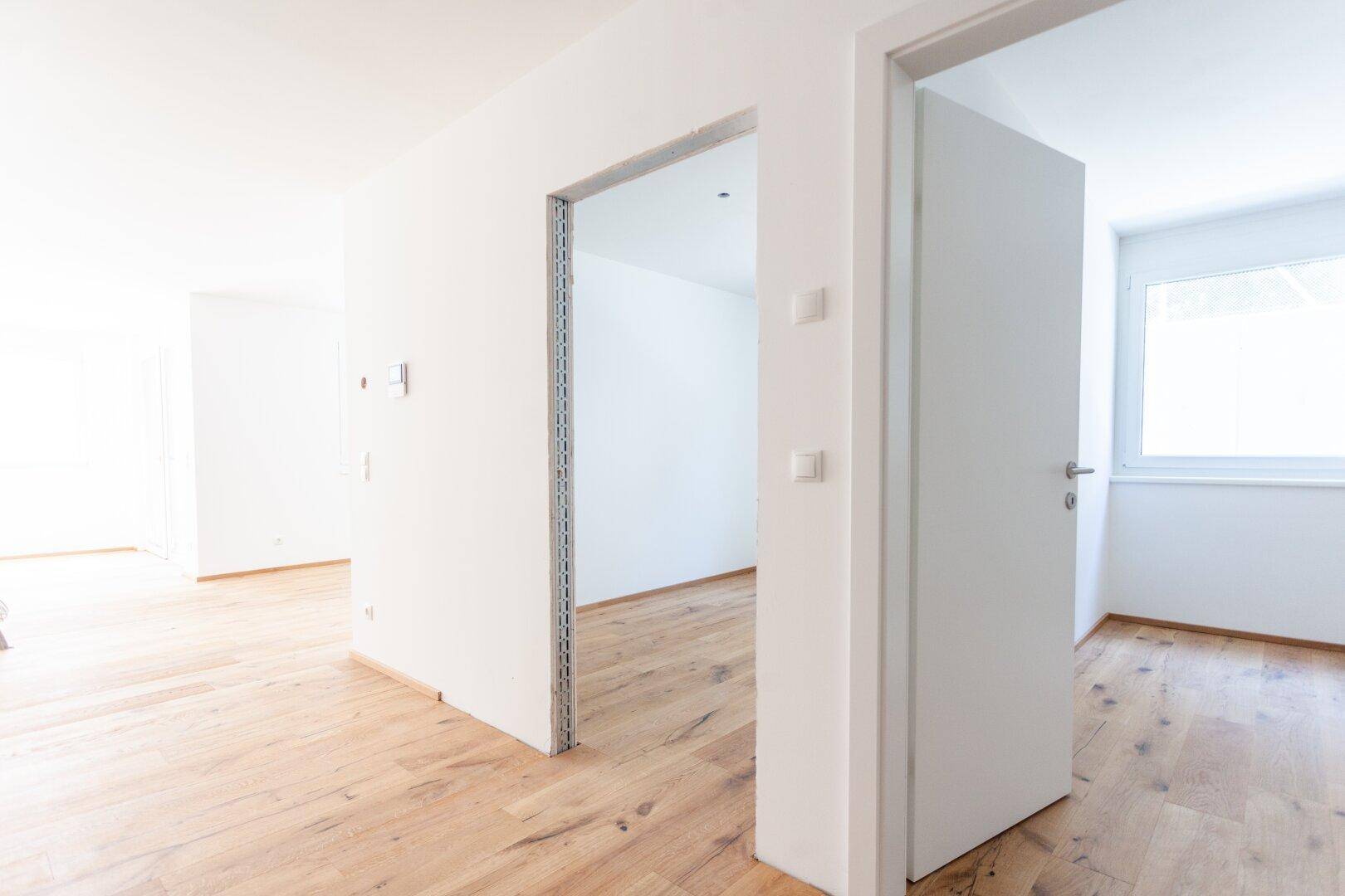 Heller Innenraum mit Holzfußboden und offener Tür zu einem weiteren Zimmer, bietet vielseitige Nutzungsmöglichkeiten.