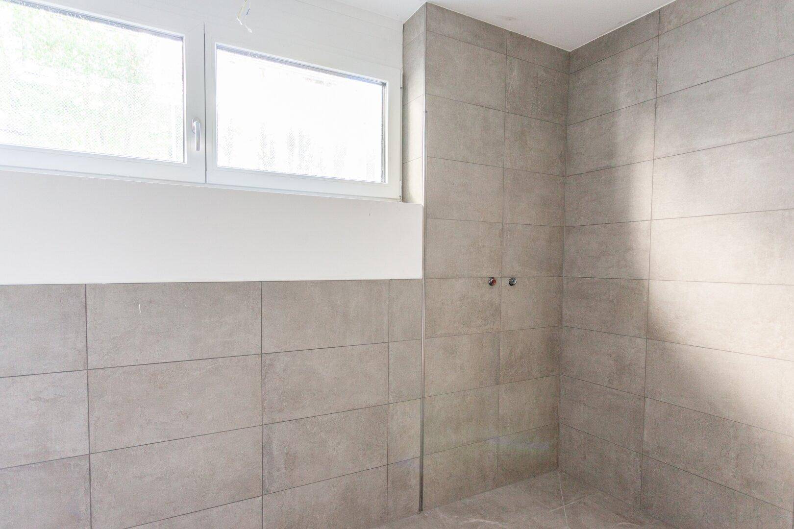 Badezimmer im Bau mit grauen Fliesen an Wänden und Boden sowie einem Fenster für Belüftung.