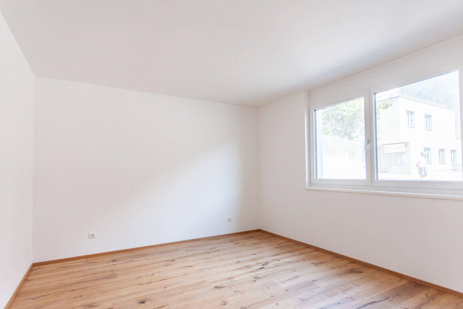 Leerer Raum mit Holzboden und einem großen Fenster, das viel natürliches Licht spendet.