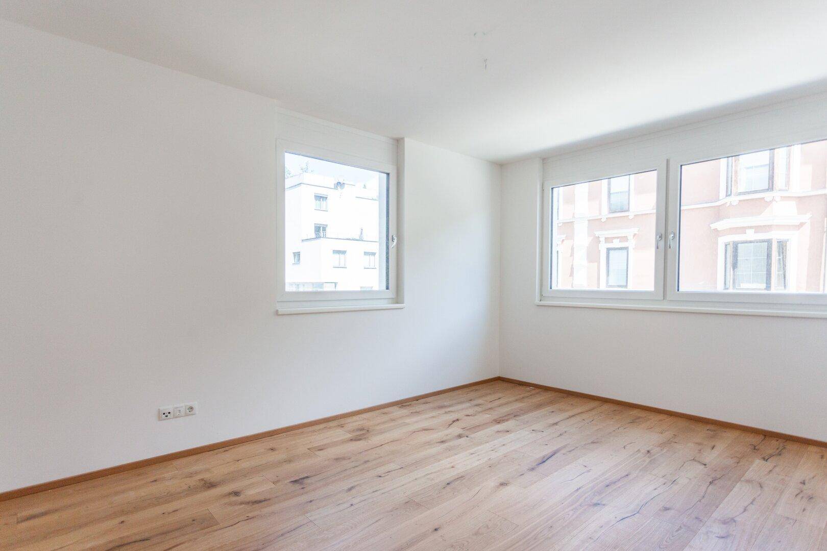 Leeres Schlafzimmer mit Holzboden und Blick auf umliegende Gebäude.