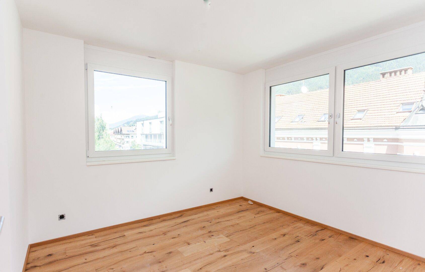 Leeres Schlafzimmer mit Holzboden, weißen Wänden und Blick auf die umliegende Bebauung und Natur.