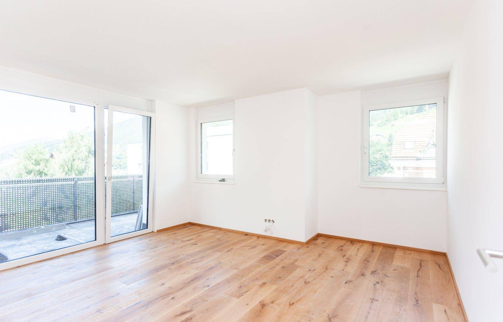 Heller Raum mit Holzboden und Fenstern, eines davon mit Zugang zu einem Balkon.