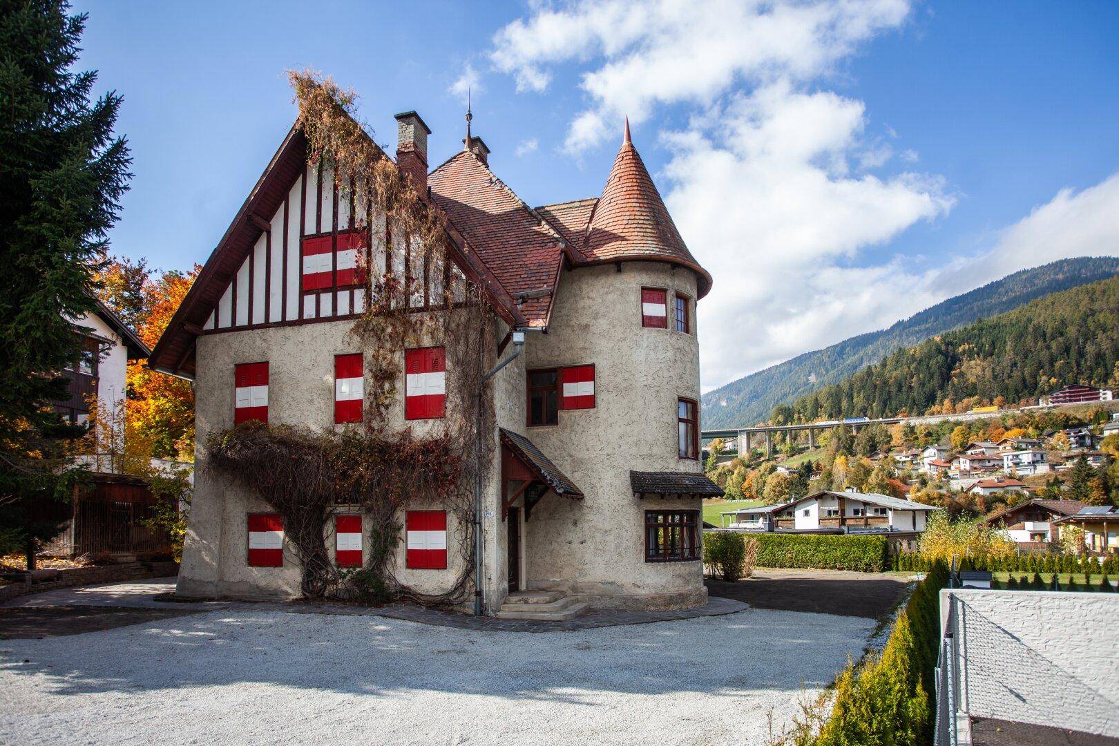Historisches Gebäude mit Turm und roten Fensterläden, umgeben von einer Kiesfläche und Bergblick.