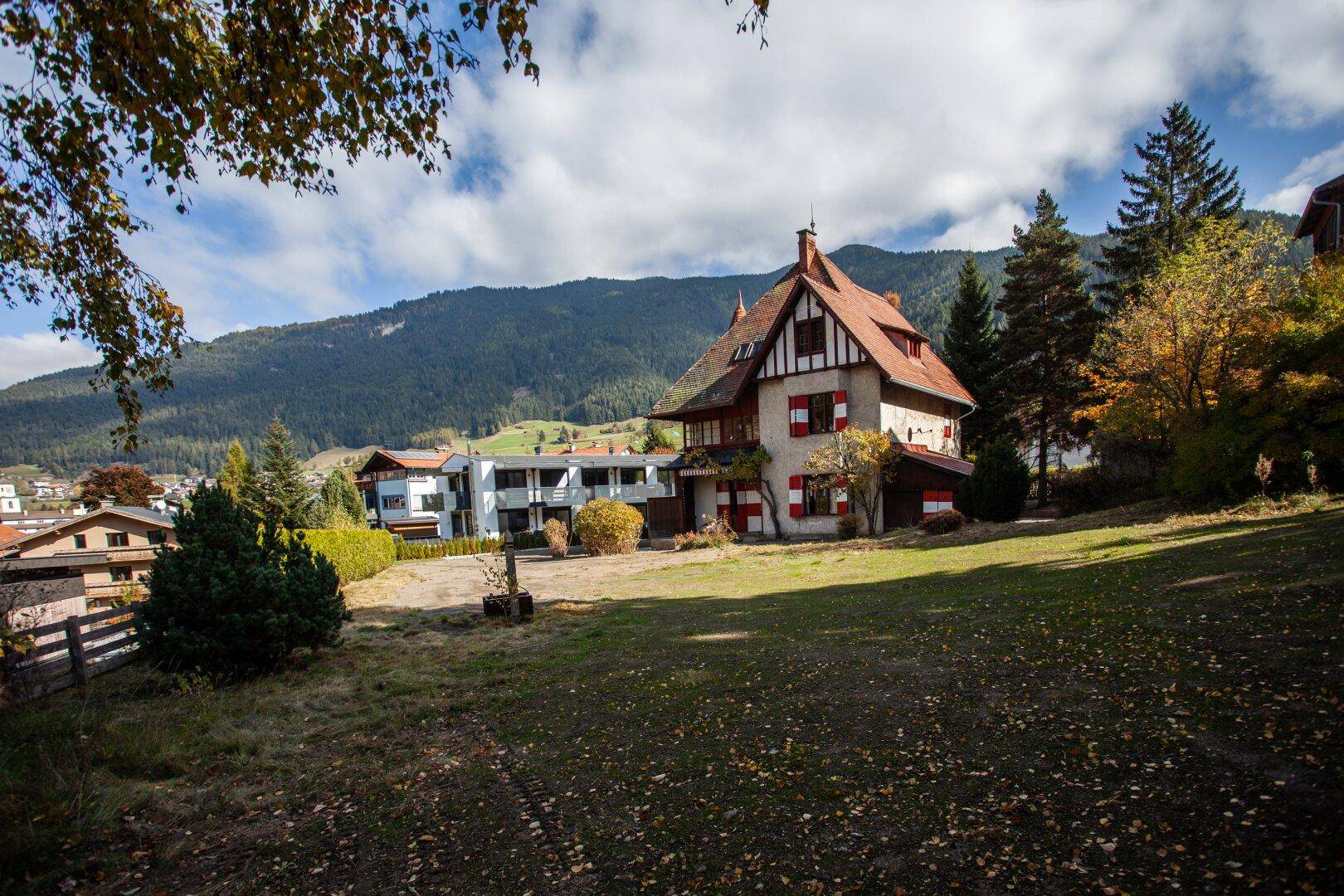 Historisches Haus mit rotem Dach und Fensterläden, umgeben von einer weitläufigen Wiese und Bergen.