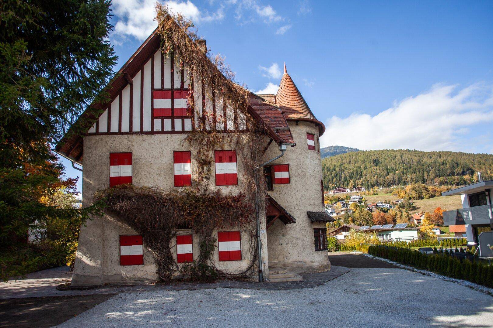 Historisches Gebäude mit markantem Turm und roten Fensterläden, umgeben von einer Kiesfläche und Bäumen.