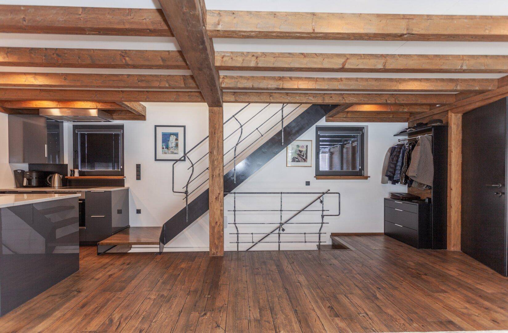 Treppenaufgang und offener Bereich mit Holzboden, moderner Küche im Hintergrund und Garderobe.