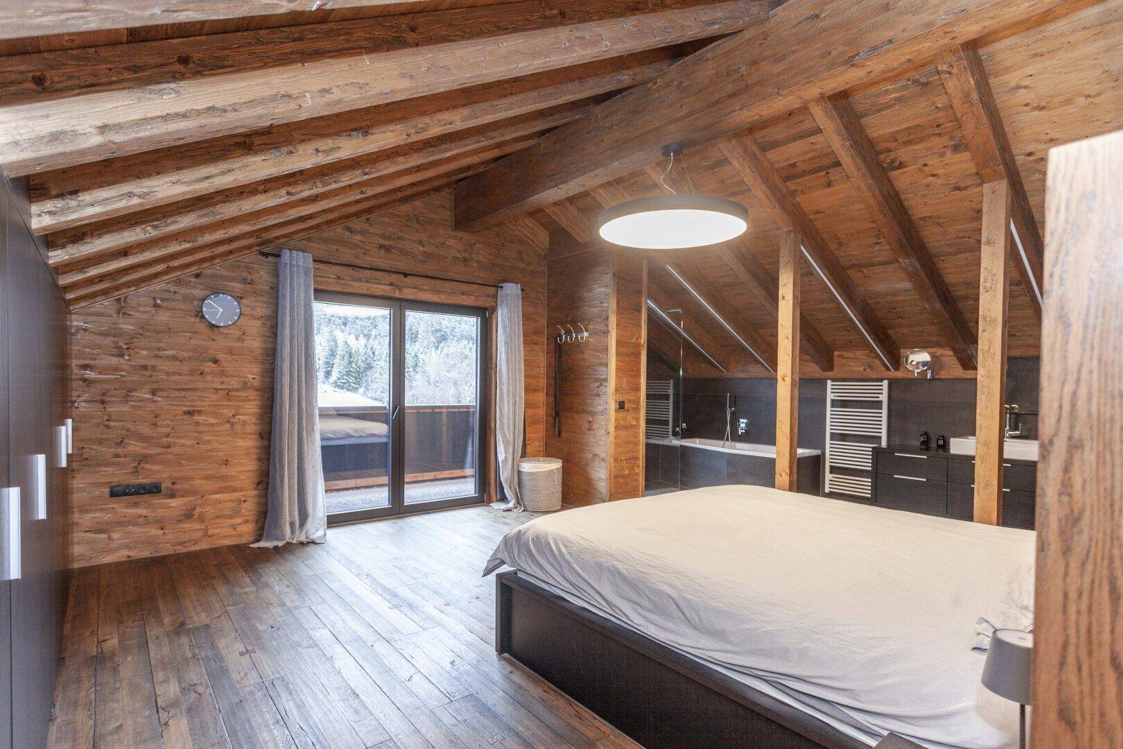 Geräumiges Schlafzimmer im Dachgeschoss mit Holzbalken, Balkonzugang und Blick auf die Natur.