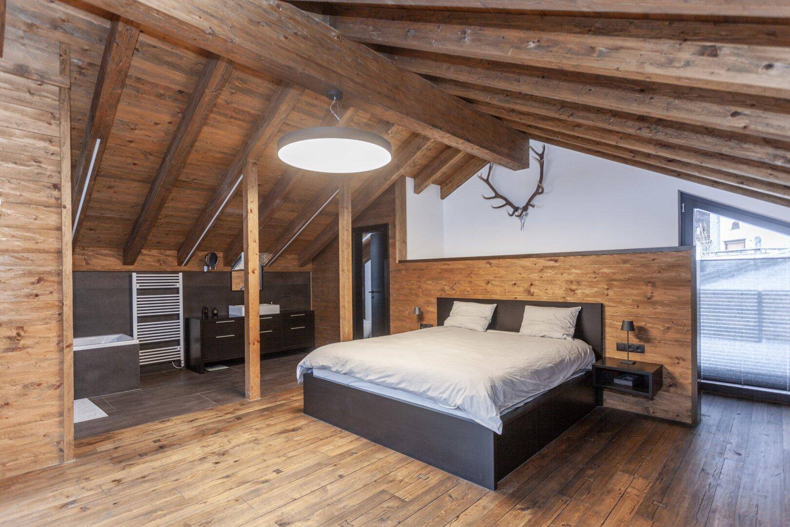Schlafzimmer im Dachgeschoss mit Holzbalken, Holzboden und integriertem Badezimmerbereich.
