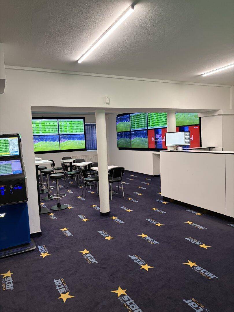 Weitläufiger Innenraum eines Wettbüros mit Thekenbereich, Spielautomaten und zahlreichen Bildschirmen für Sportwetten.