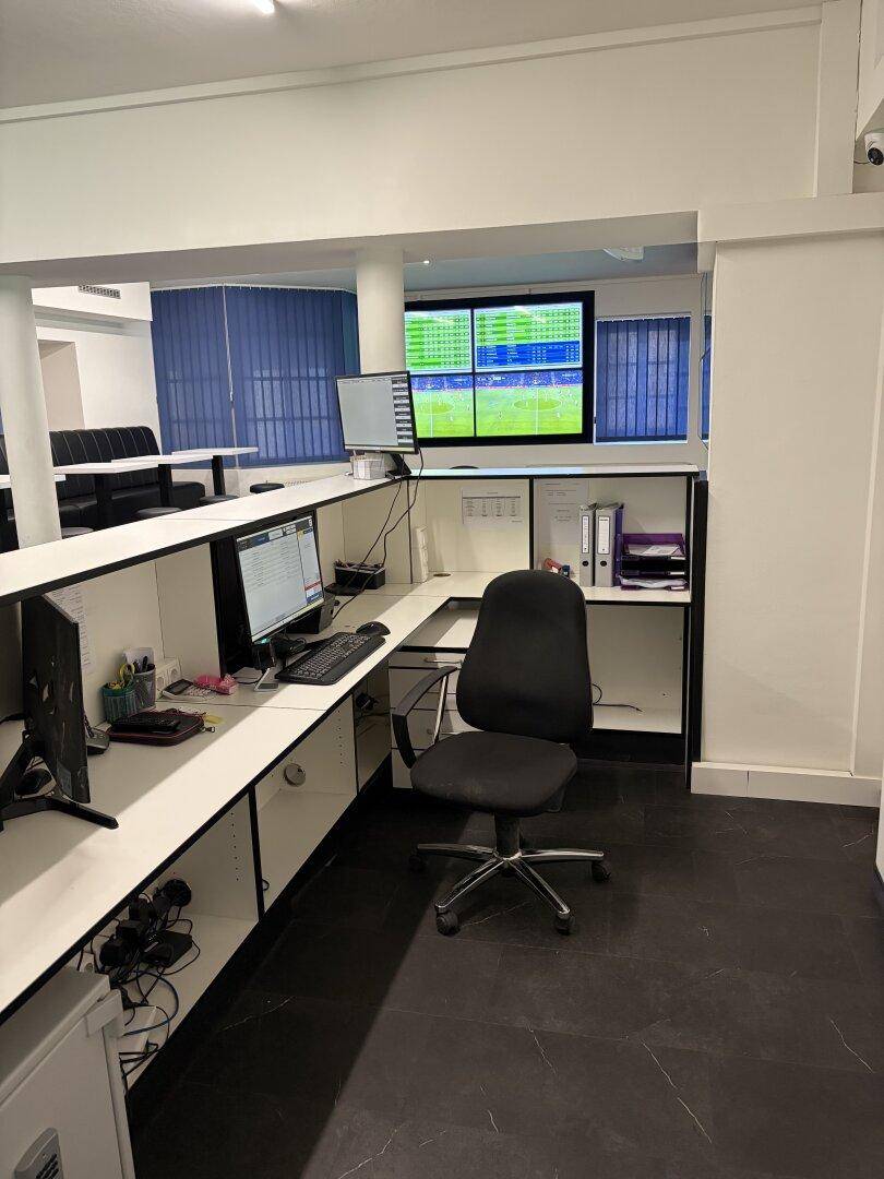 Bürobereich hinter der Theke mit Schreibtisch, Bürostuhl, Computermonitoren und Akten, alles in einem funktionalen Arbeitsumfeld.