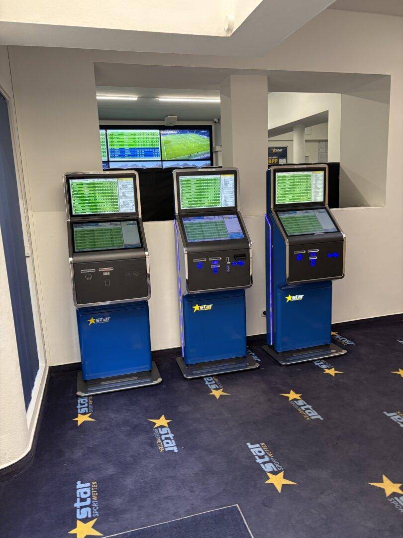 Drei moderne Wettterminals in einer Reihe, bereit zur Nutzung, mit blauen Jalousien im Hintergrund.