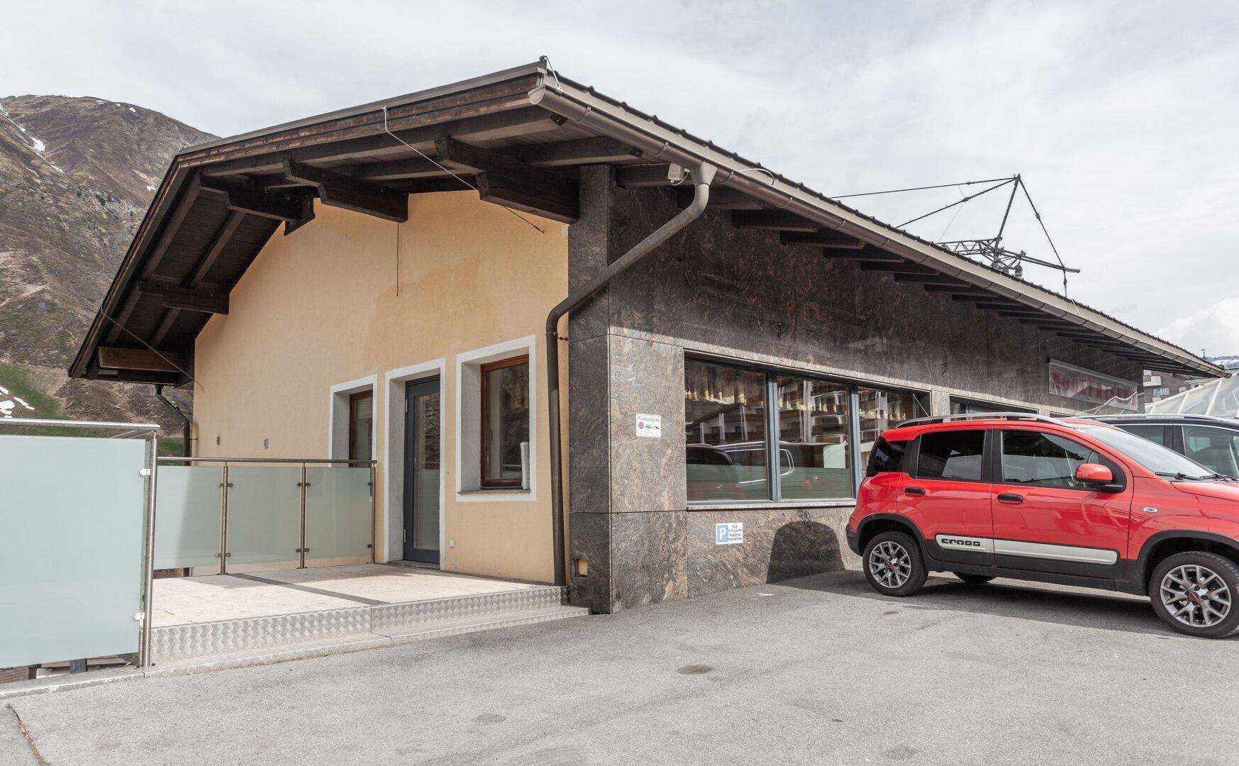 Seitenansicht eines Gebäudes mit Parkplatz, Terrasse und Blick auf die umliegende Berglandschaft.
