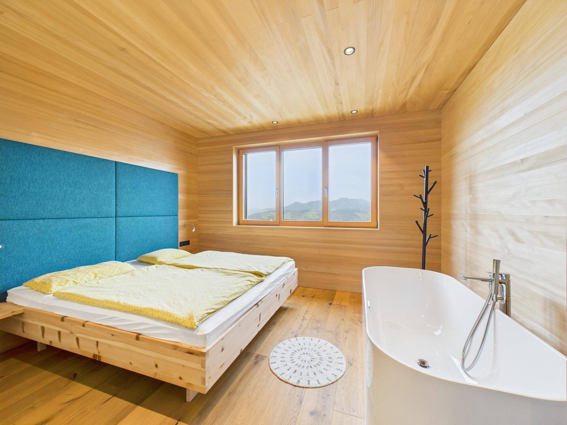 Schlafzimmer mit Holzwänden und -decke, Doppelbett und freistehender Badewanne mit Bergblick.
