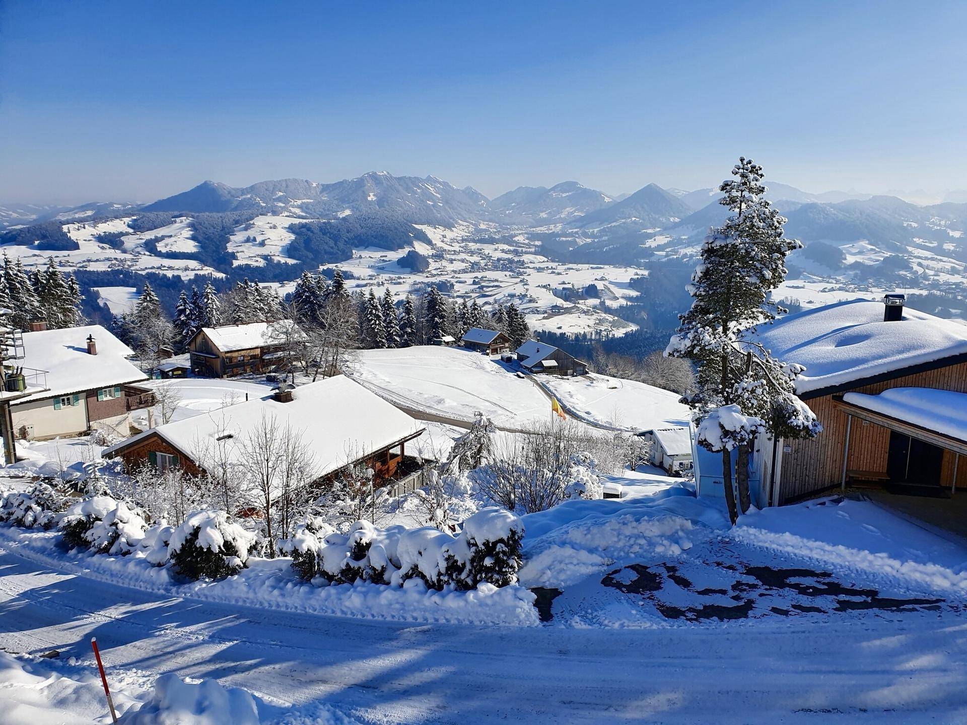 Weitläufige Winterlandschaft mit verschneiten Häusern und Bergen unter blauem Himmel.