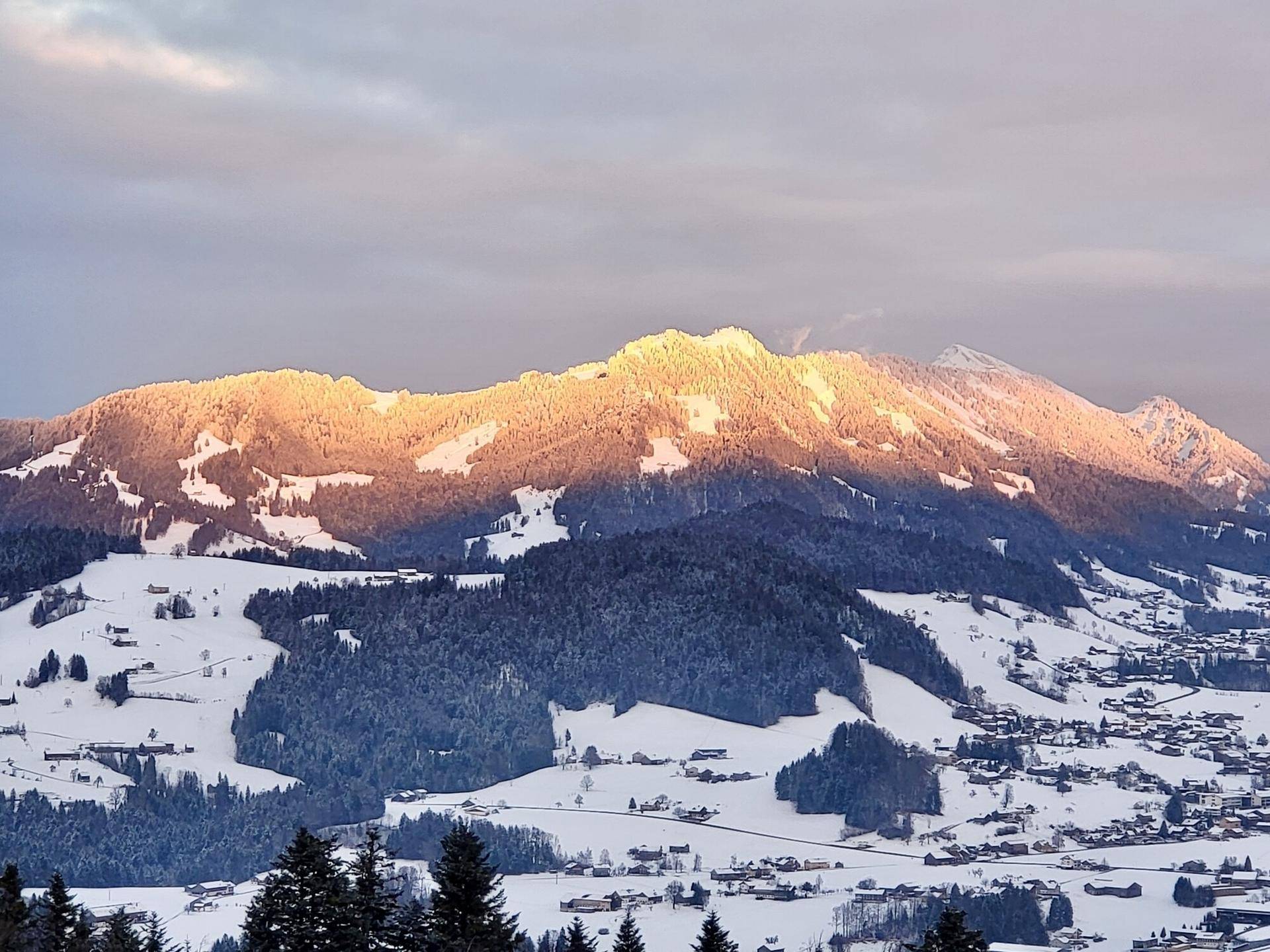 Sonnenuntergang über schneebedeckten Bergen und einem Tal mit kleinen Siedlungen.