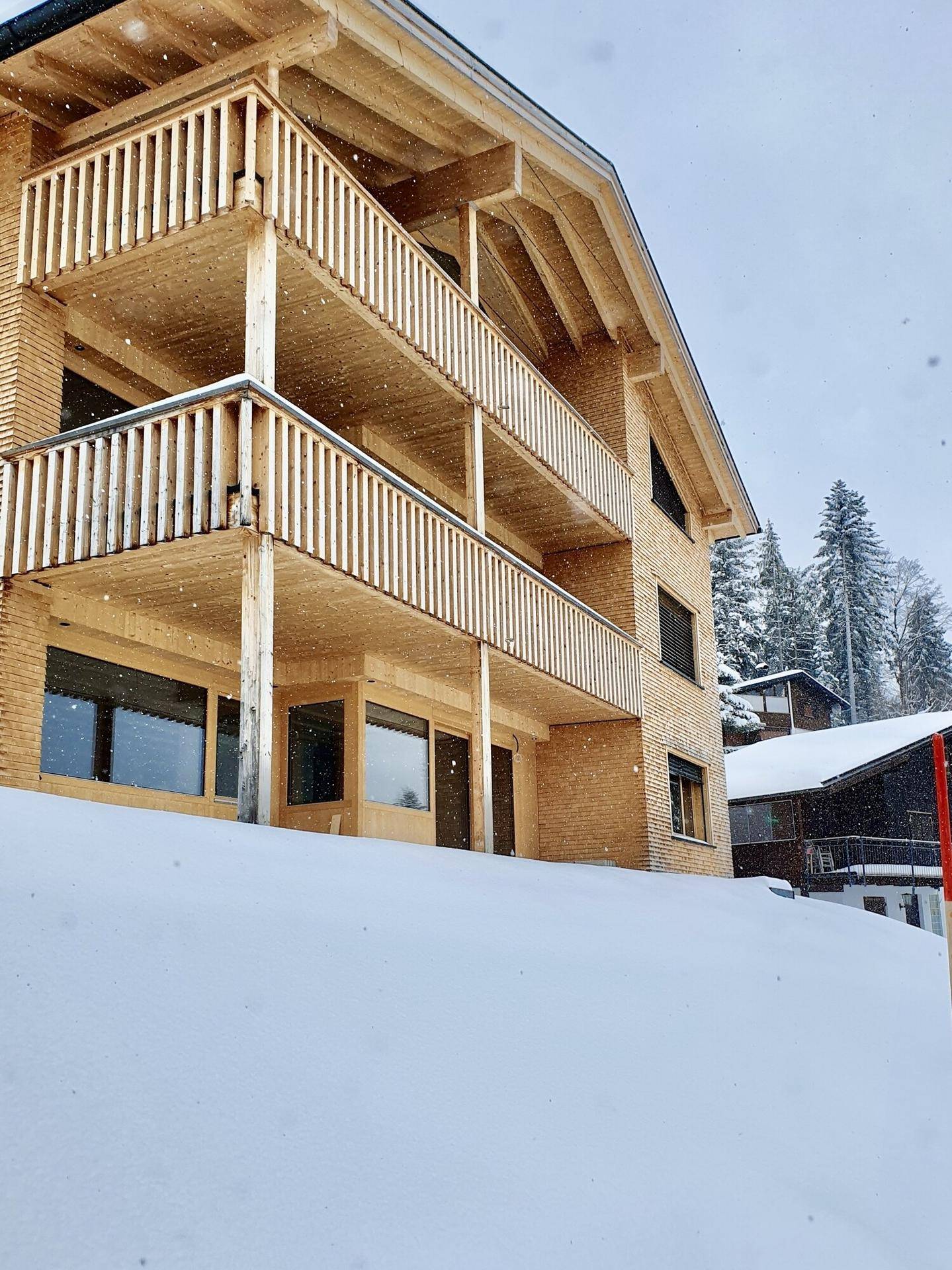 Neubau mit Holzfassade und mehreren Balkonen, umgeben von Schnee und Bäumen.