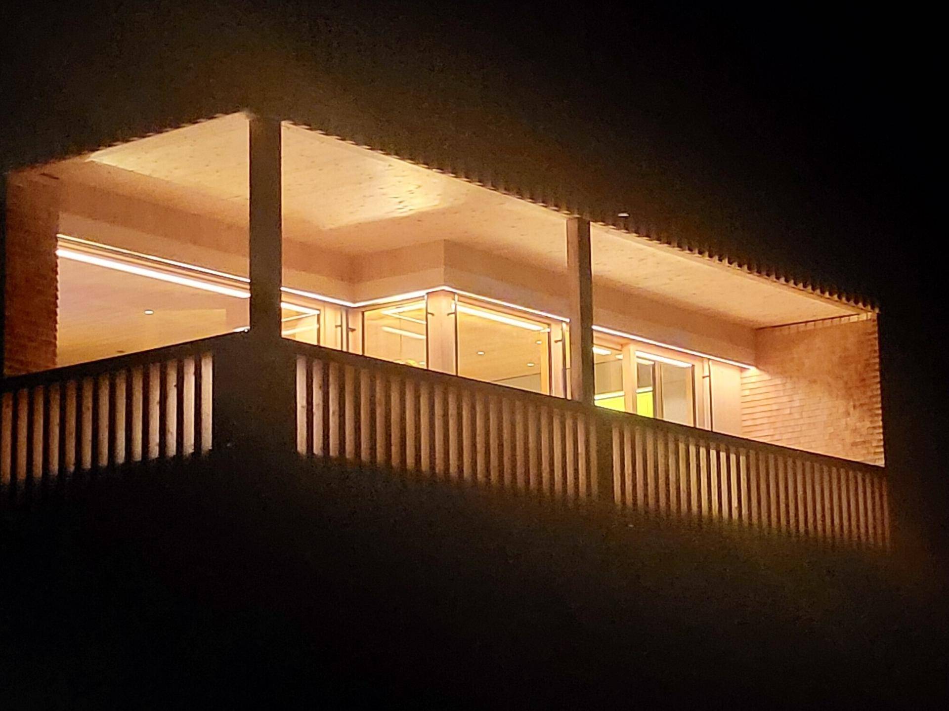 Beleuchtete Holzfassade eines Gebäudes bei Nacht mit sichtbaren Balkonen und Fenstern.