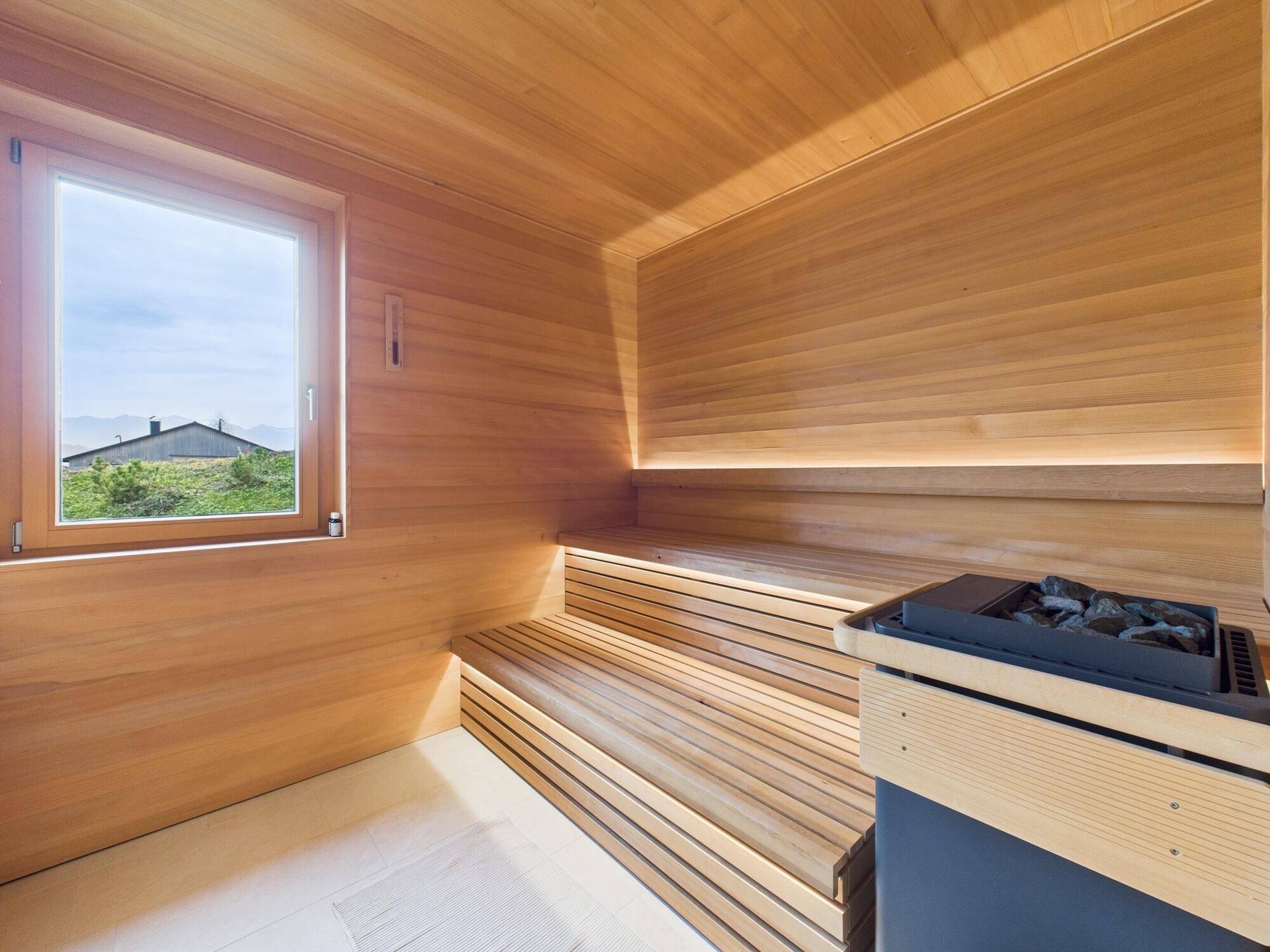Holzverkleidete Sauna mit mehrstufigen Bänken, Saunaofen und Fenster mit Ausblick.