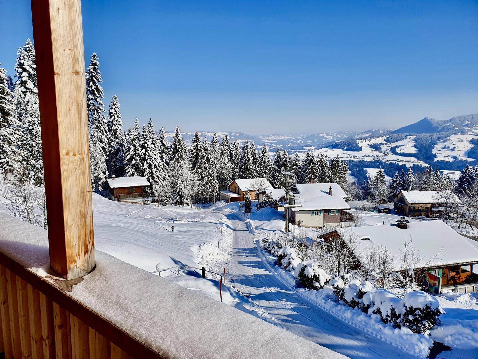 Winterliche Berglandschaft mit verschneiten Häusern und Tannenbäumen, gesehen von einem Balkon.