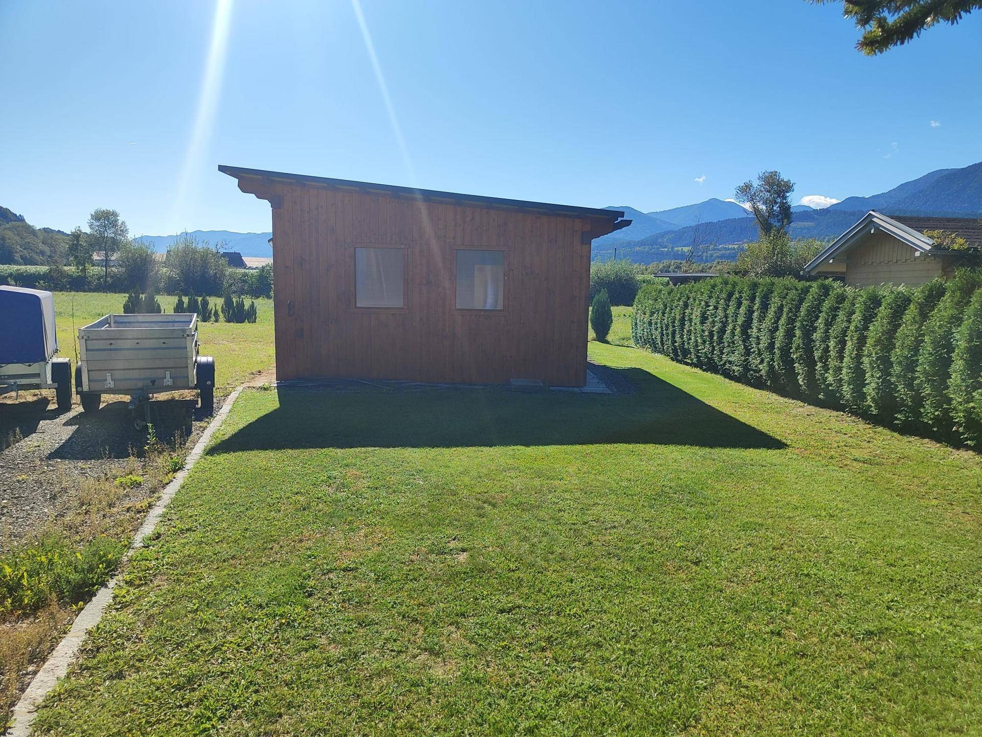 Kleines Holzhaus im Garten mit gepflegtem Rasen und einer hohen Hecke im Hintergrund an einem sonnigen Tag.