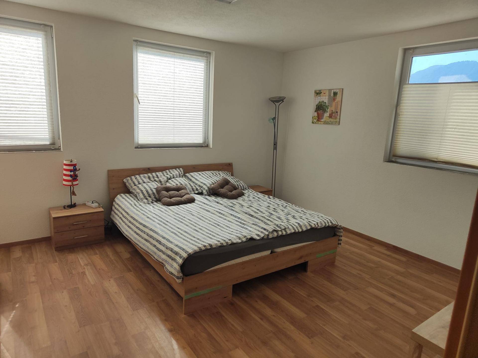 Helles Schlafzimmer mit Doppelbett, Holzfußboden und zwei großen Fenstern mit Jalousien.