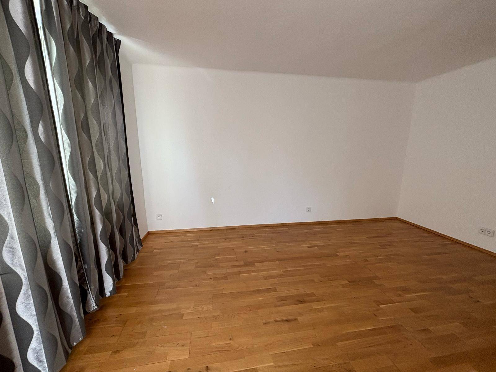 Leeres Zimmer mit Parkettboden und dunklen Vorhängen vor dem Fenster.