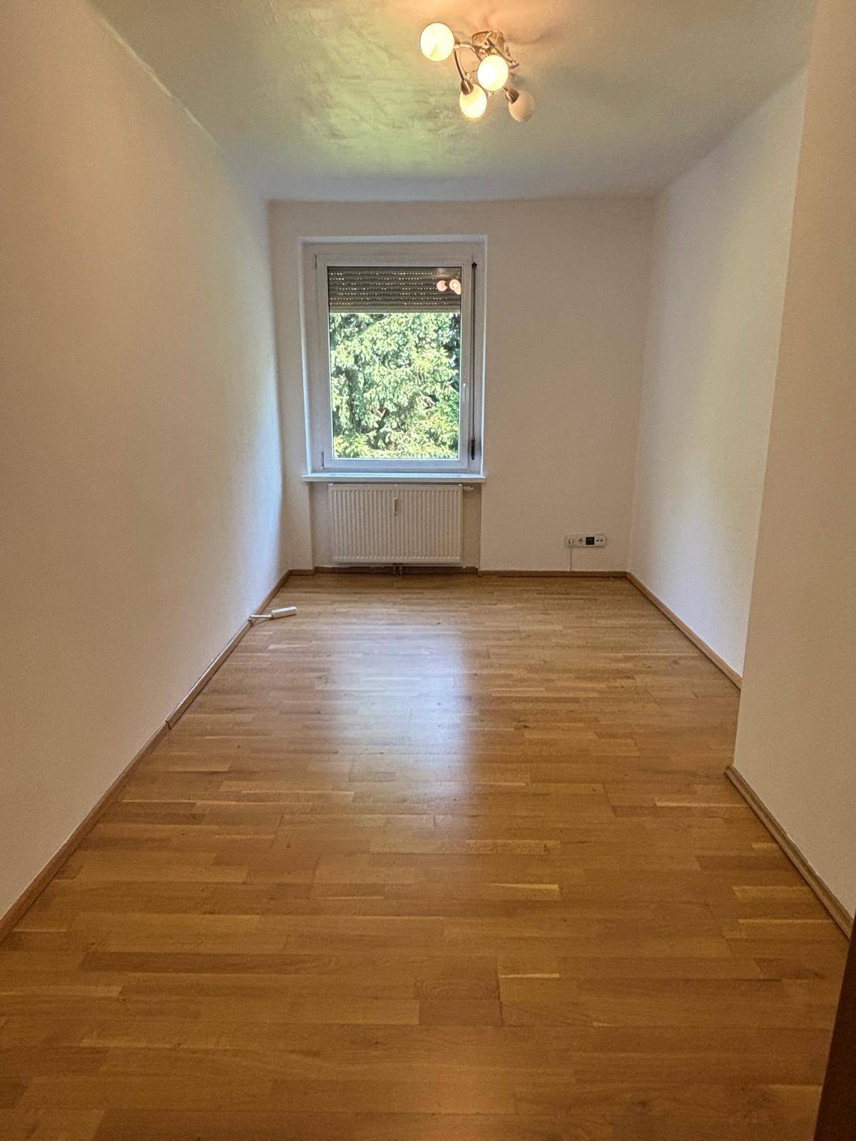 Leeres Zimmer mit Parkettboden und Fenster mit Blick ins Grüne.