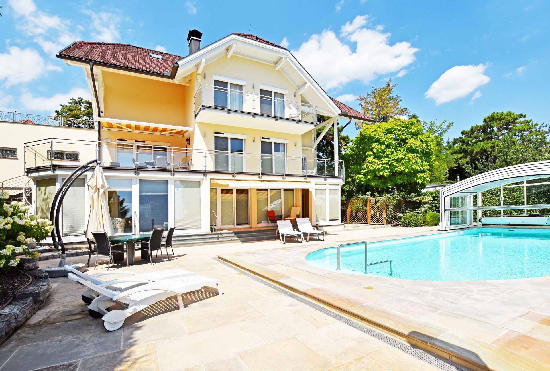 Sonnige Terrasse mit Poolbereich und Liegestühlen vor der eleganten Hausfassade und Garten.