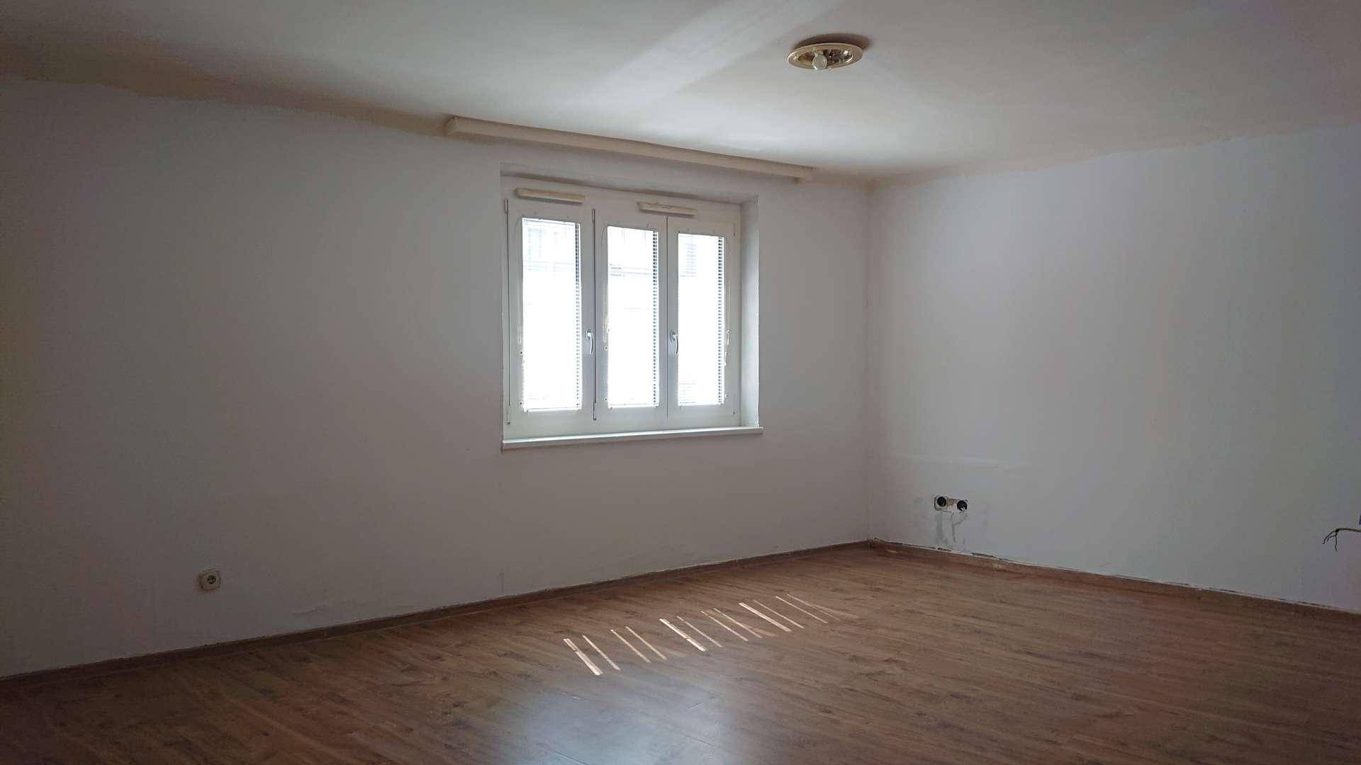 Leerer Raum mit Holzboden und einem großen Fenster, das viel Tageslicht hereinlässt.