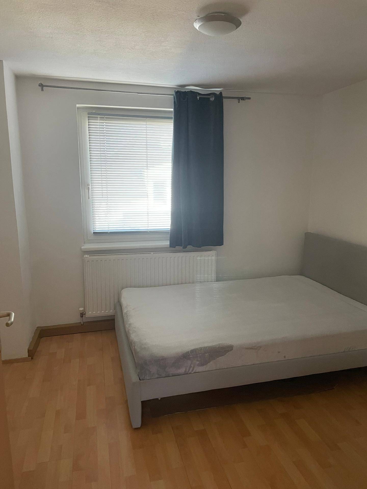 Schlafzimmer mit Laminatboden, einem Fenster und einem Bett mit einfacher Matratze.