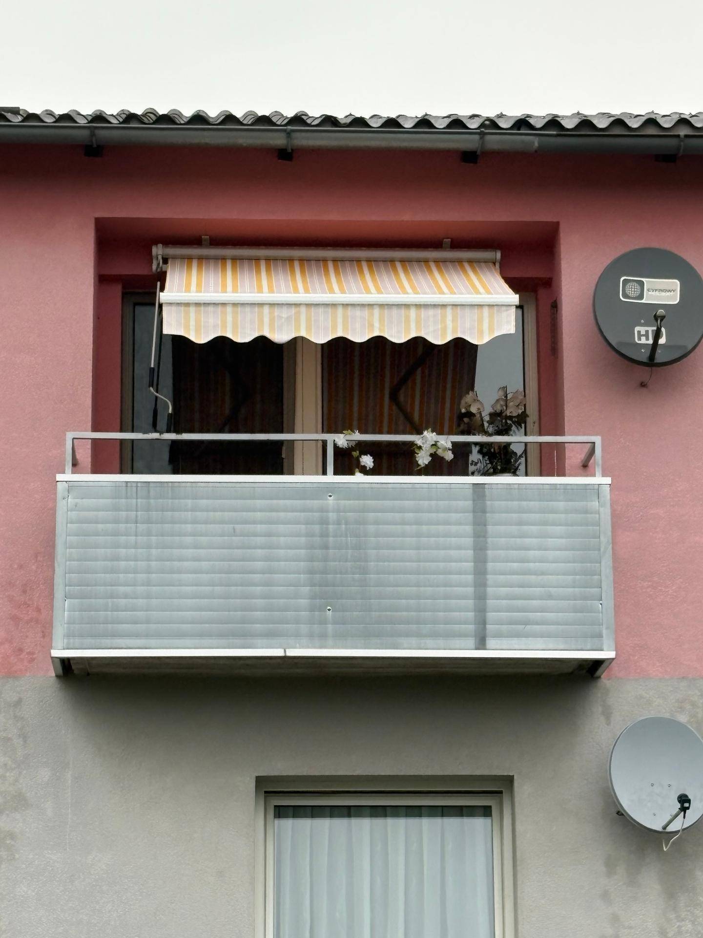 Kleiner Balkon an einer rosafarbenen Fassade mit gestreifter Markise und Satellitenschüsseln.