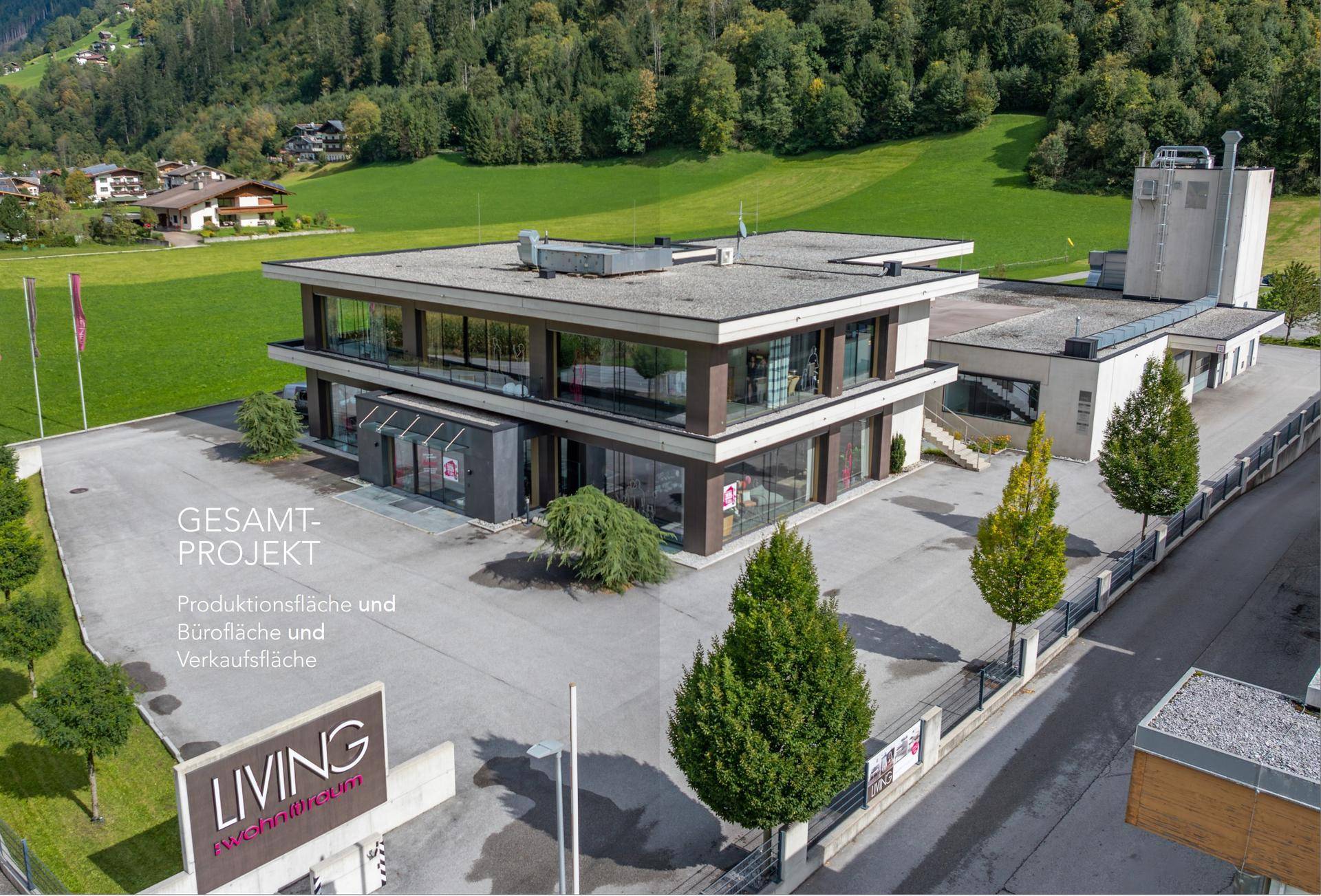 Luftaufnahme des modernen Gewerbegebäudes mit großzügigen Parkplätzen, umgeben von grünen Wiesen und einer malerischen Bergkulisse.