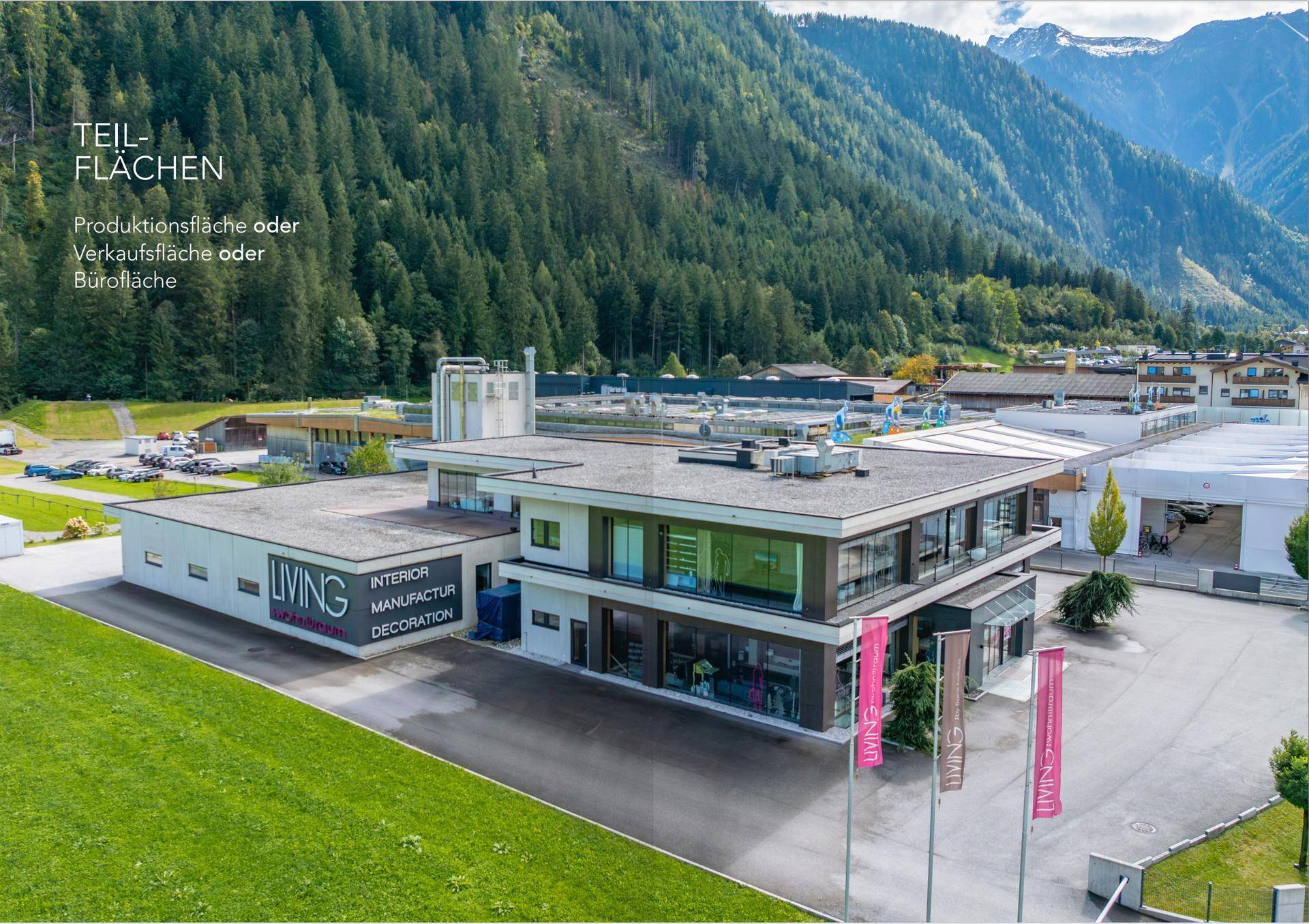 Luftaufnahme eines modernen Gewerbegebäudes mit Flachdach, umgeben von grünen Wiesen und einer beeindruckenden Berglandschaft.