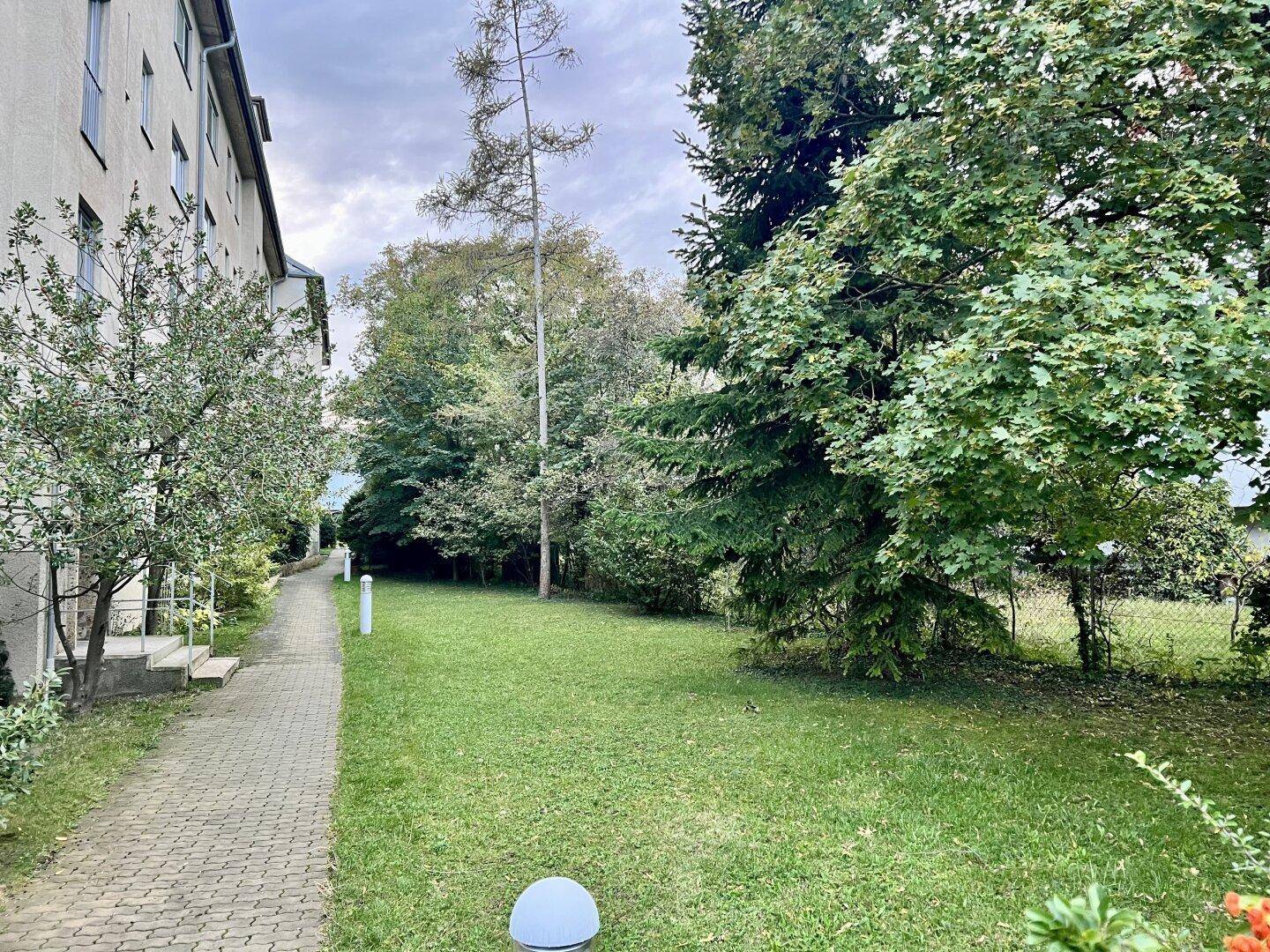 Gepflasterter Weg führt entlang eines Gebäudes zu einer grünen Rasenfläche mit Bäumen.