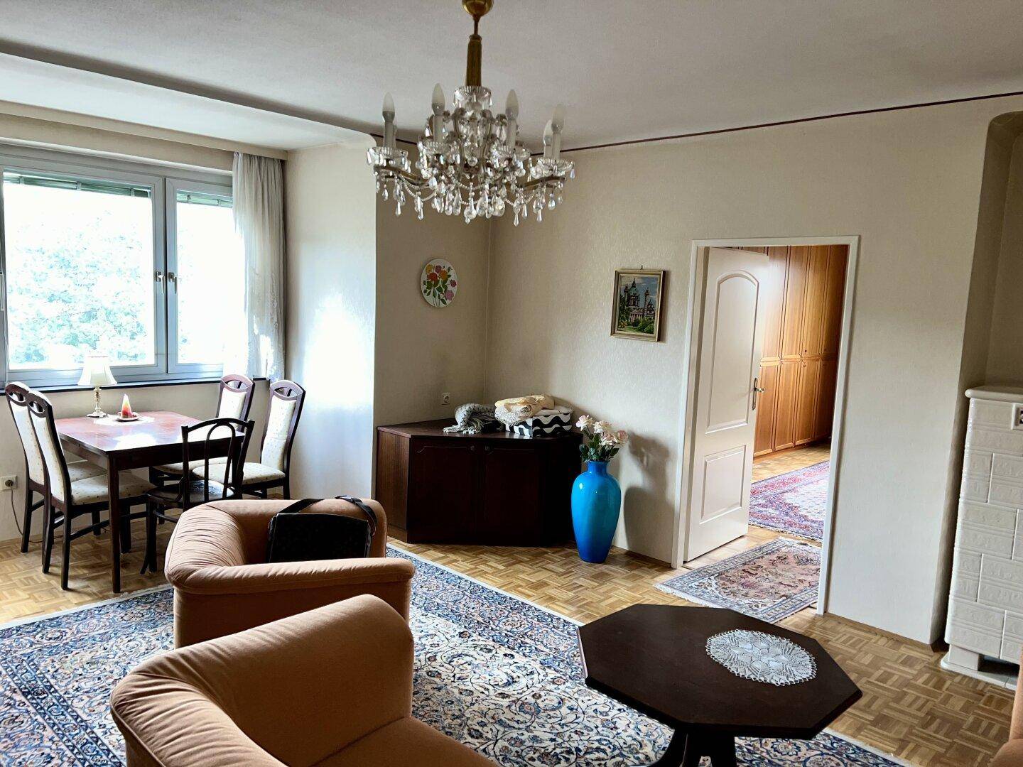 Wohn- und Essbereich mit Kronleuchter, Teppich und Blick auf einen weiteren Raum.