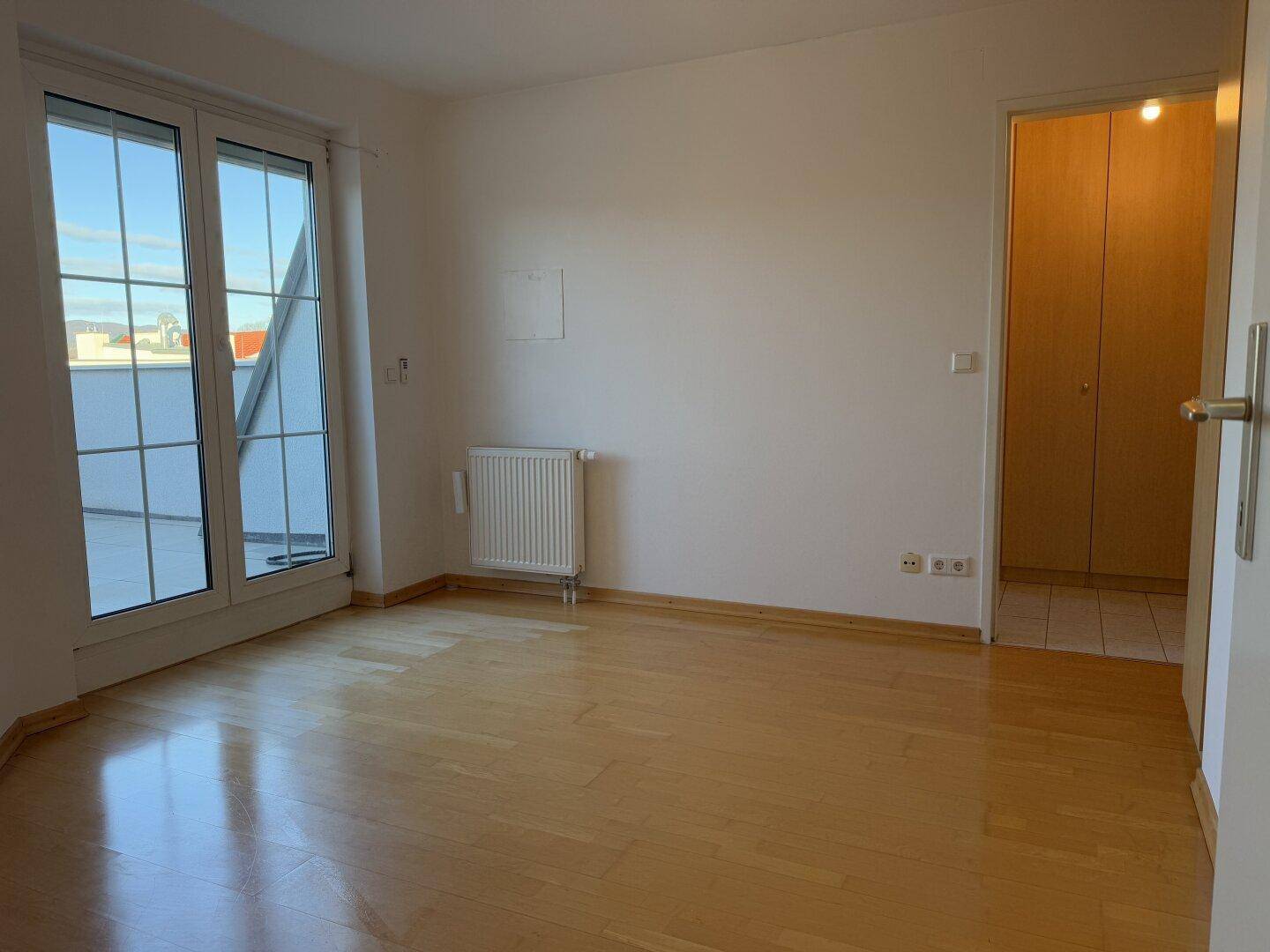 Heller Raum mit Parkettboden und großen Fenstern, die Zugang zu einem Balkon bieten.