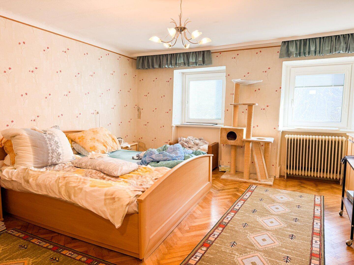 Schlafzimmer mit Holzbett, gemusterter Tapete und zwei Fenstern für natürliches Licht.